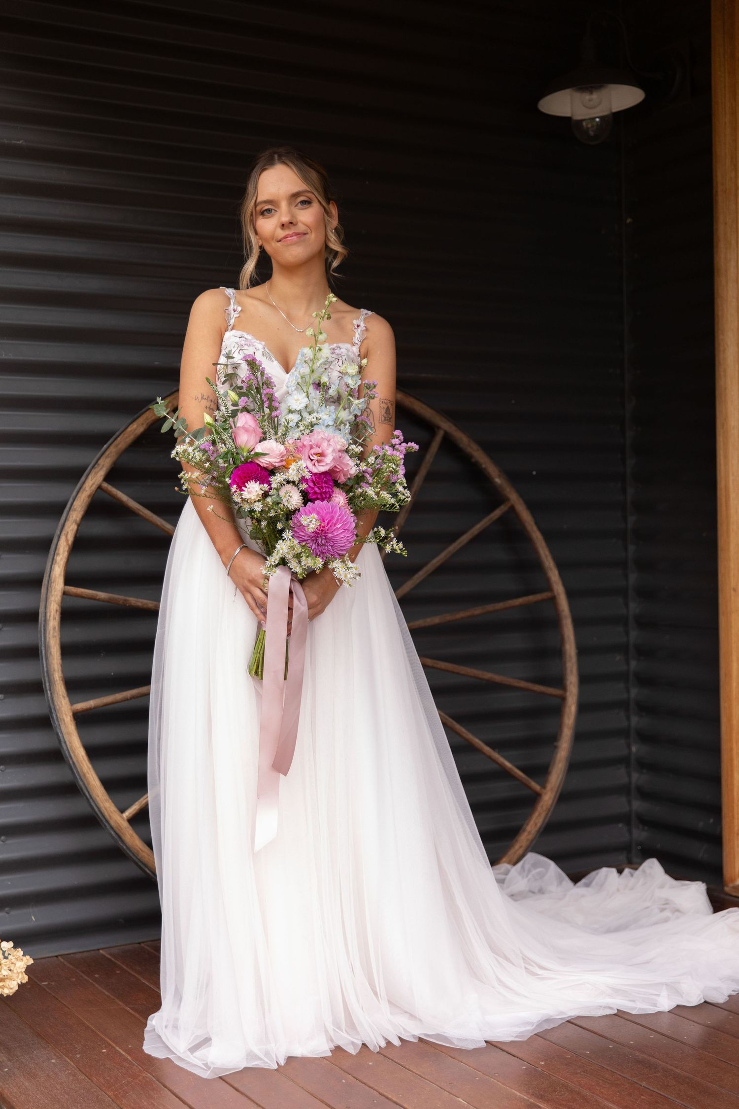 Bride in white flowy gown holds vibrant pink bouquet, posing before rustic wooden wheel backdrop.