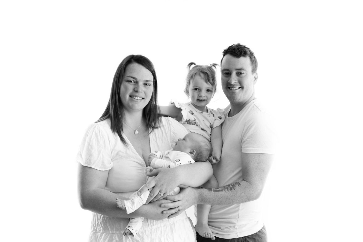 A young family in matching white outfits poses together for a heartwarming black and white portrait with their newborn baby.