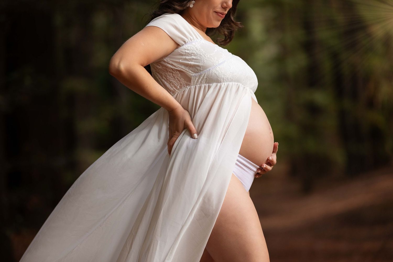 Expecting mother in flowing white dress poses in nature during maternity photoshoot with forest backdrop.
