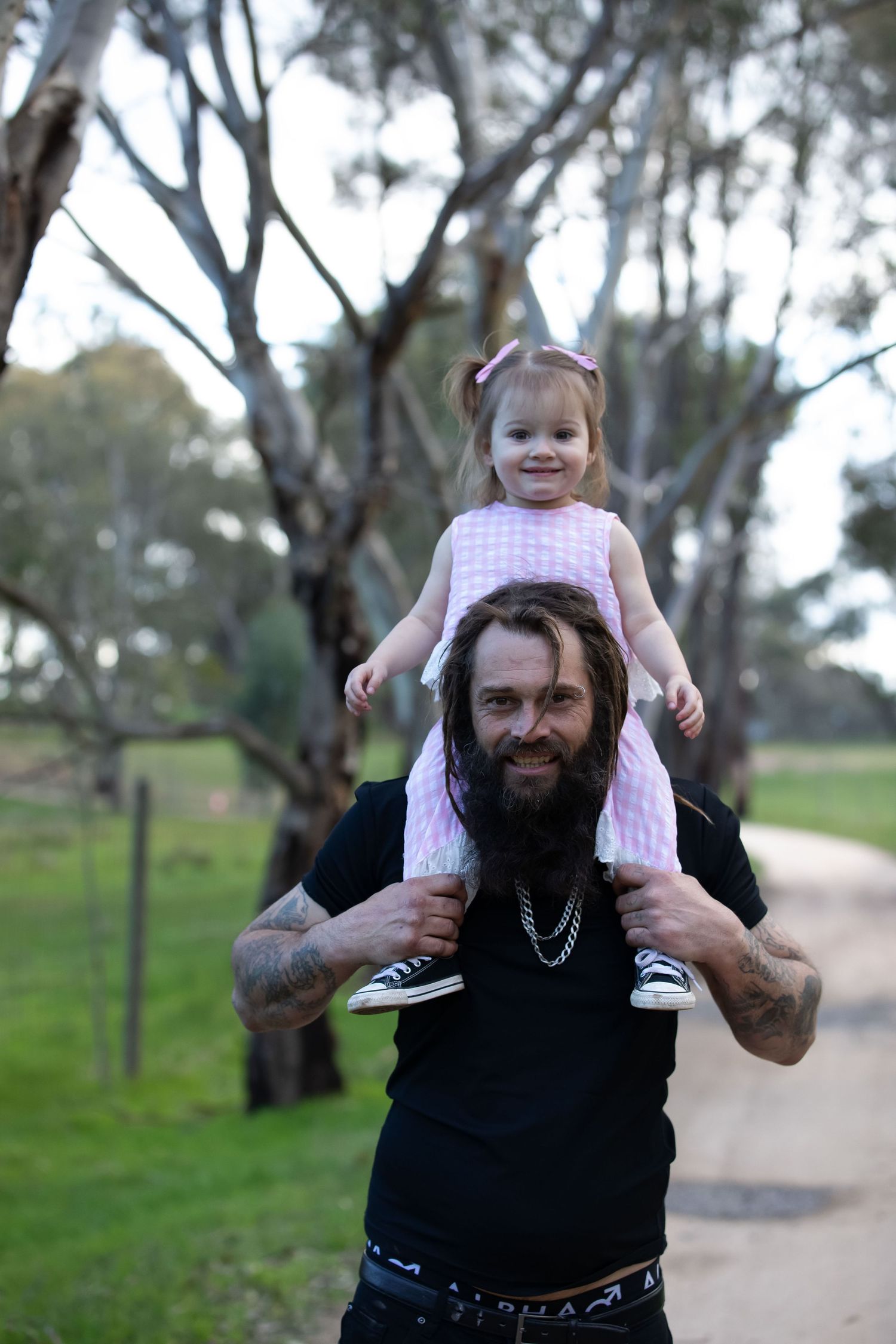 Bearded tattooed man with young girl sitting on his shoulders outdoors among eucalyptus trees on a dirt path.