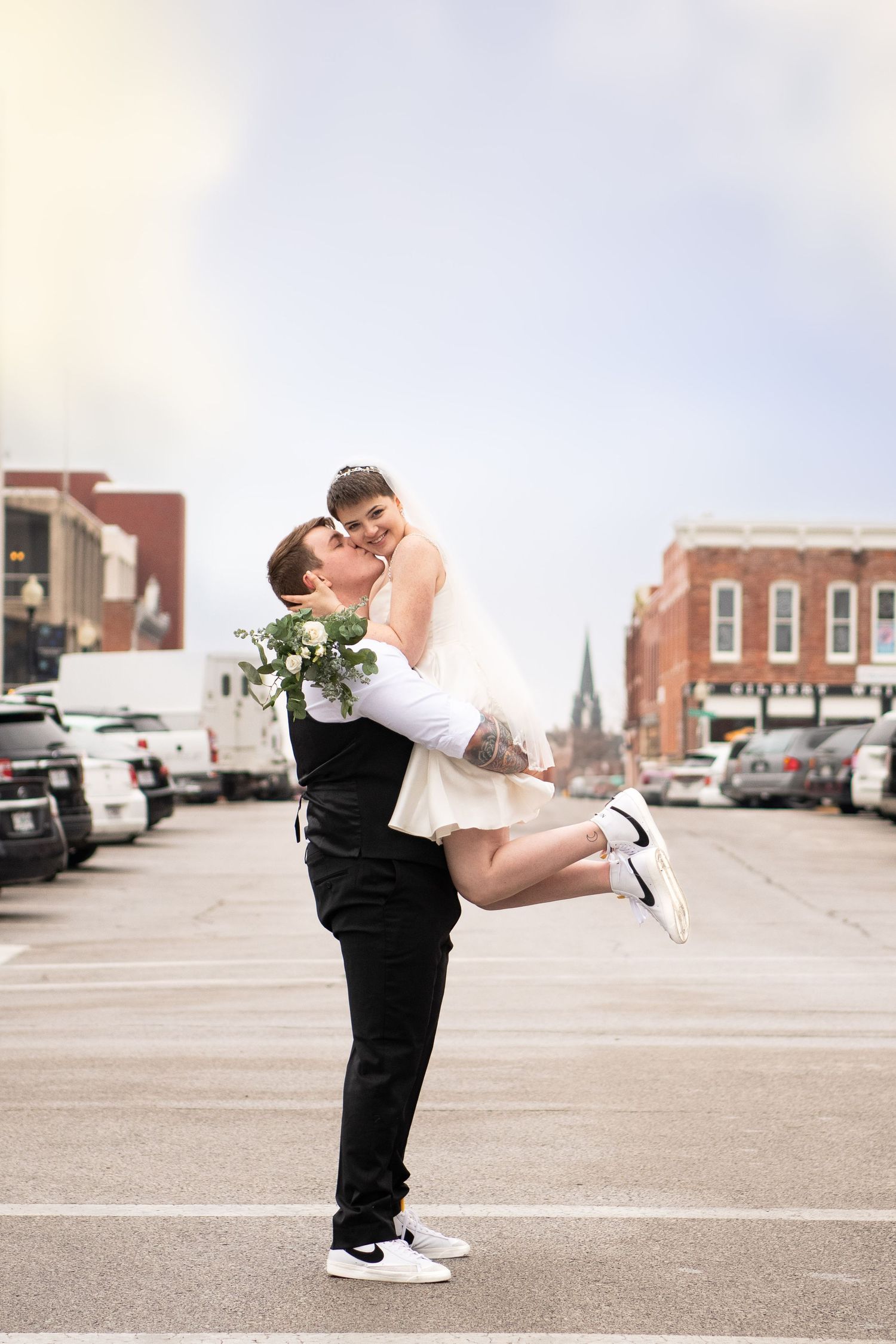 Romantic couple embraces in urban setting with bride being lifted in white dress against city backdrop.