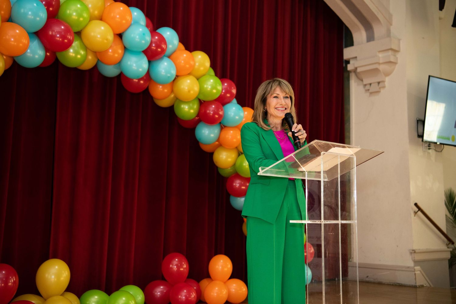 Speaker at a podium delivers remarks beneath a colorful balloon arch display at an indoor event.
