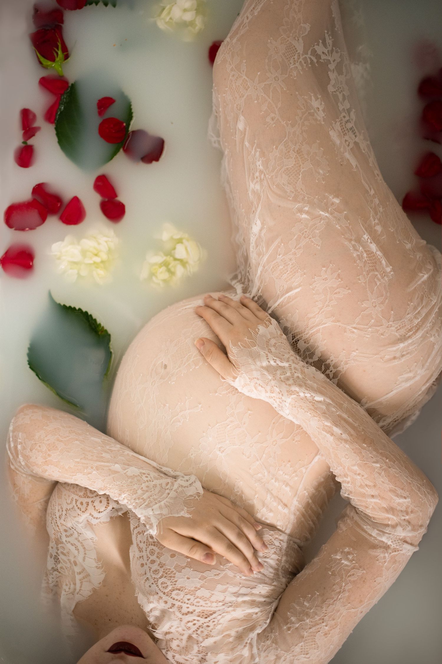 Artistic maternity milk bath photo with rose petals and lace overlay.