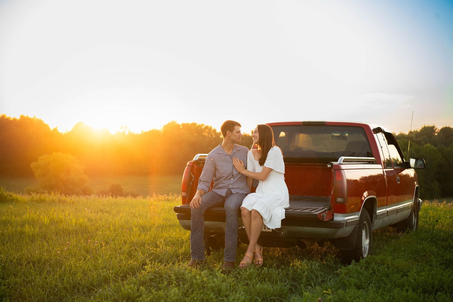 Couple sitting on pickup truck tailgate at sunset in rural field setting.