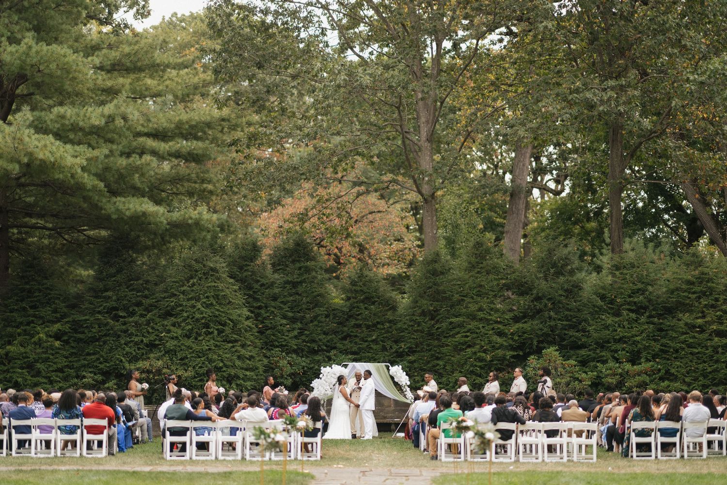 An outdoor wedding ceremony with white chairs arranged on a lawn surrounded by tall evergreen trees.