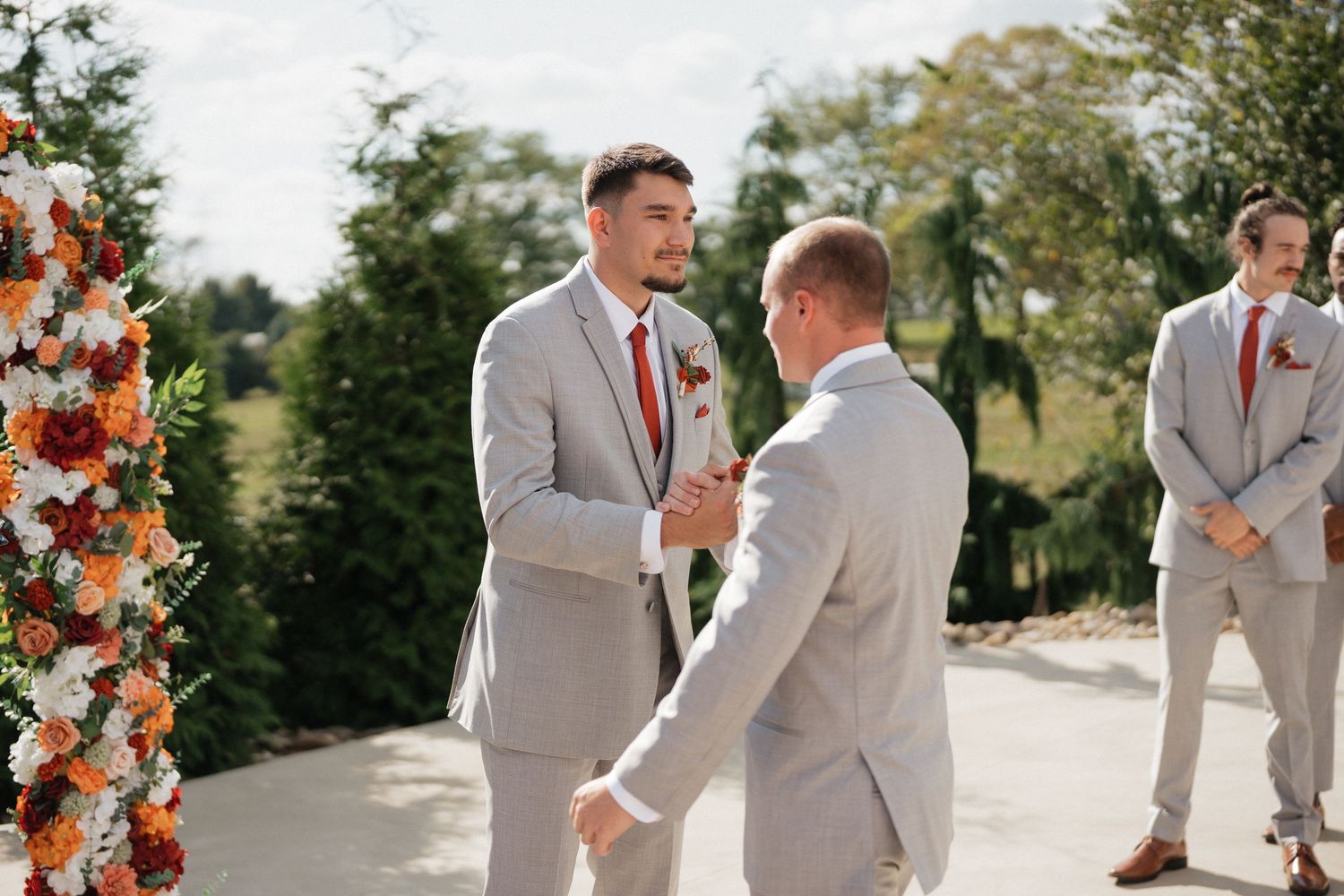 Wedding party in gray suits and orange bouquets pose outdoors on a sunny day.