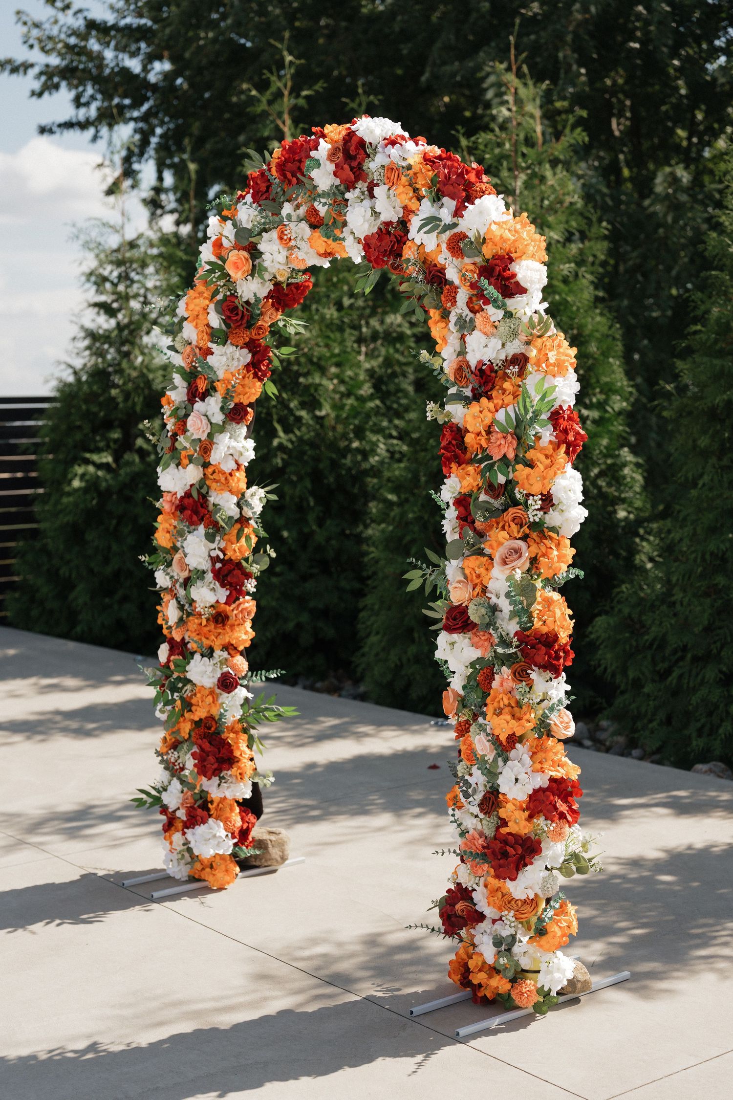 Floral wedding arch with orange, white and red flowers arranged in a curved design against a natural outdoor backdrop.