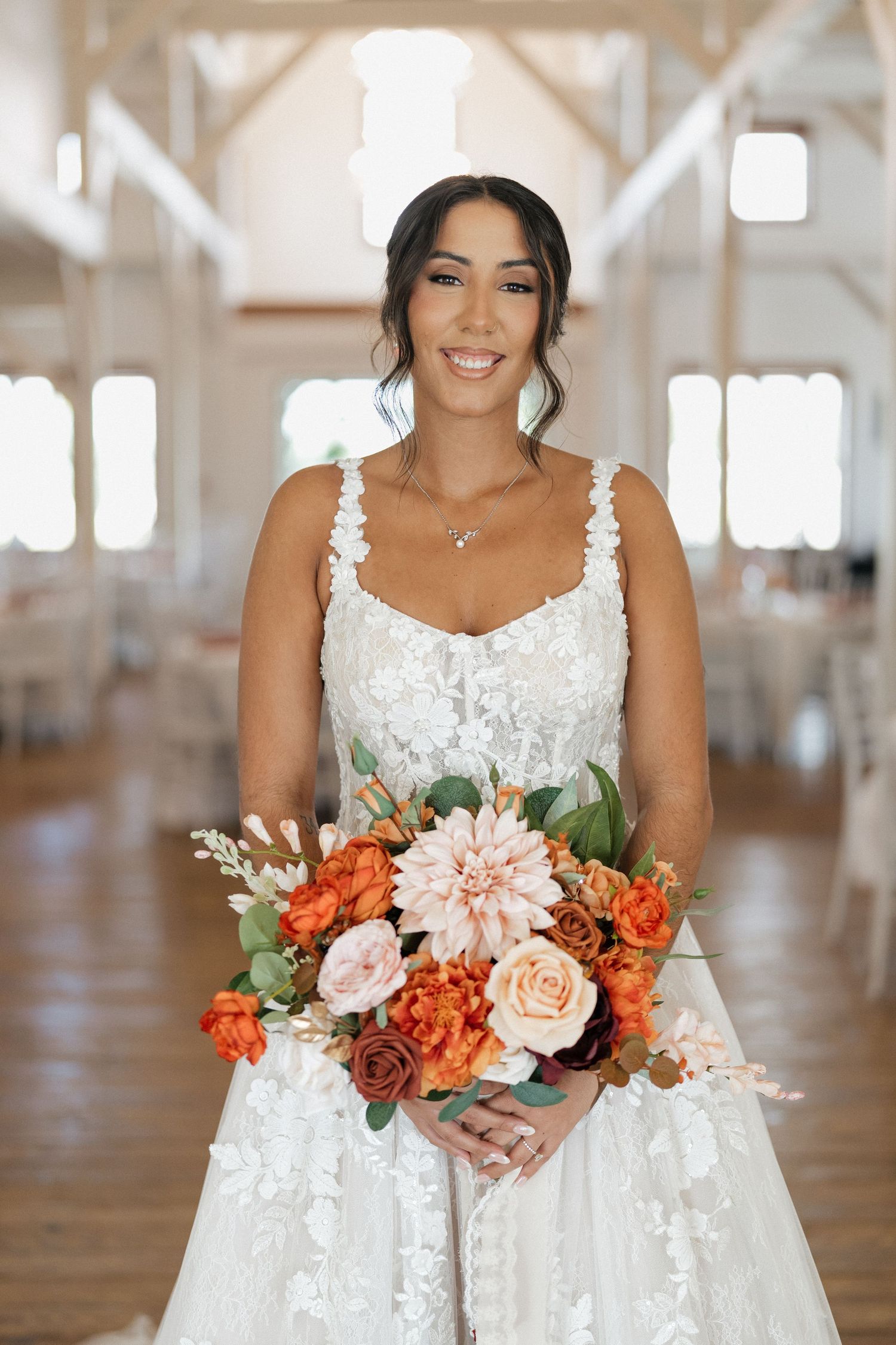 A bride in a white lace dress holds a vibrant orange and pink floral bouquet in a bright indoor venue.