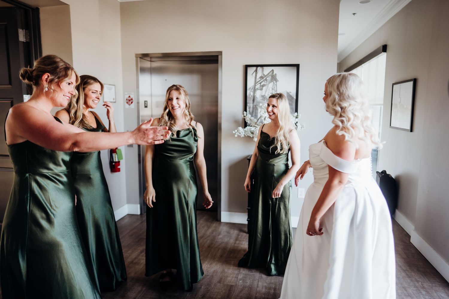 Wedding party in green dresses and white gown gather for candid moments in a bright indoor venue.