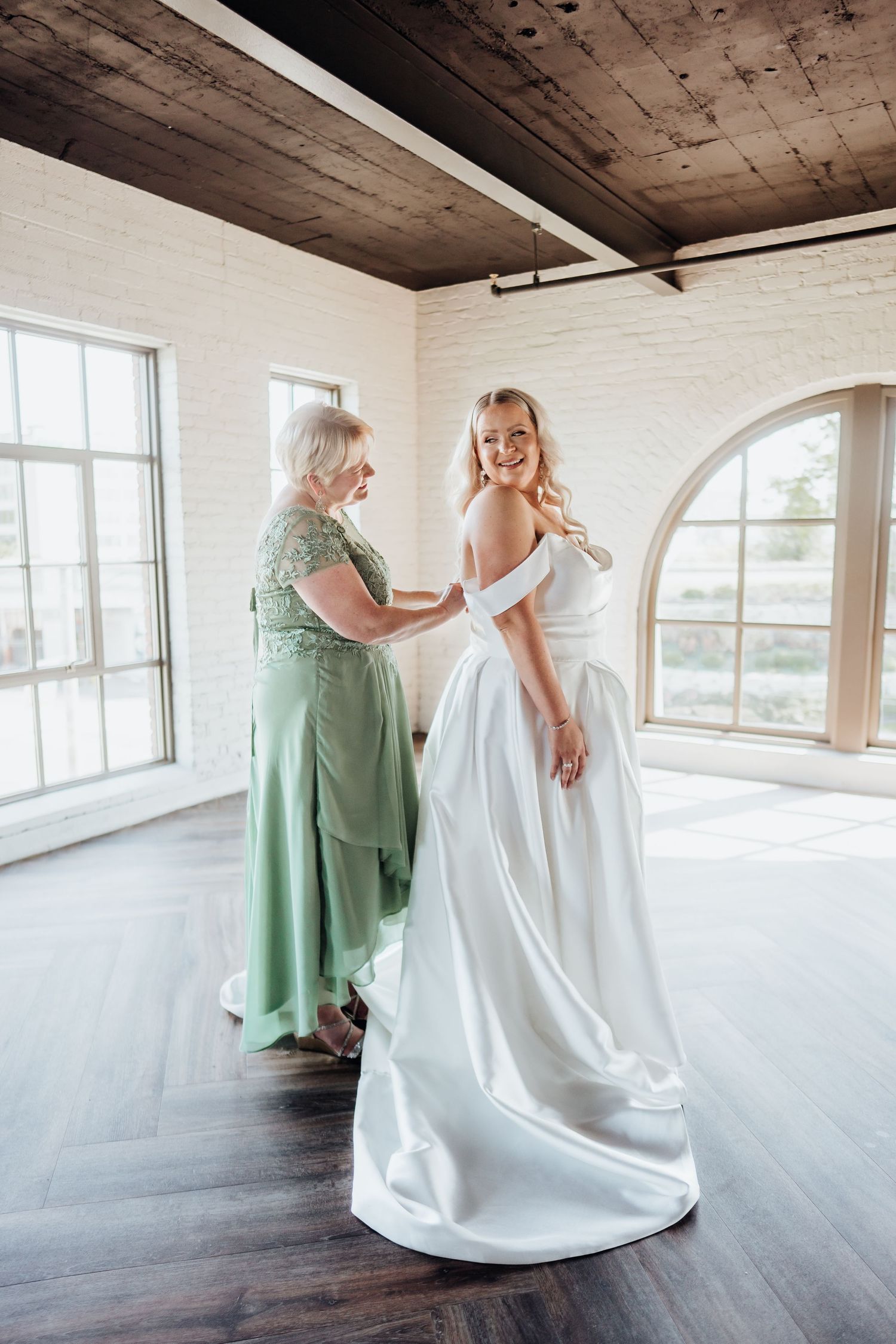 A woman in a white wedding gown gets help with her dress in a bright room with wooden ceiling and arched windows.