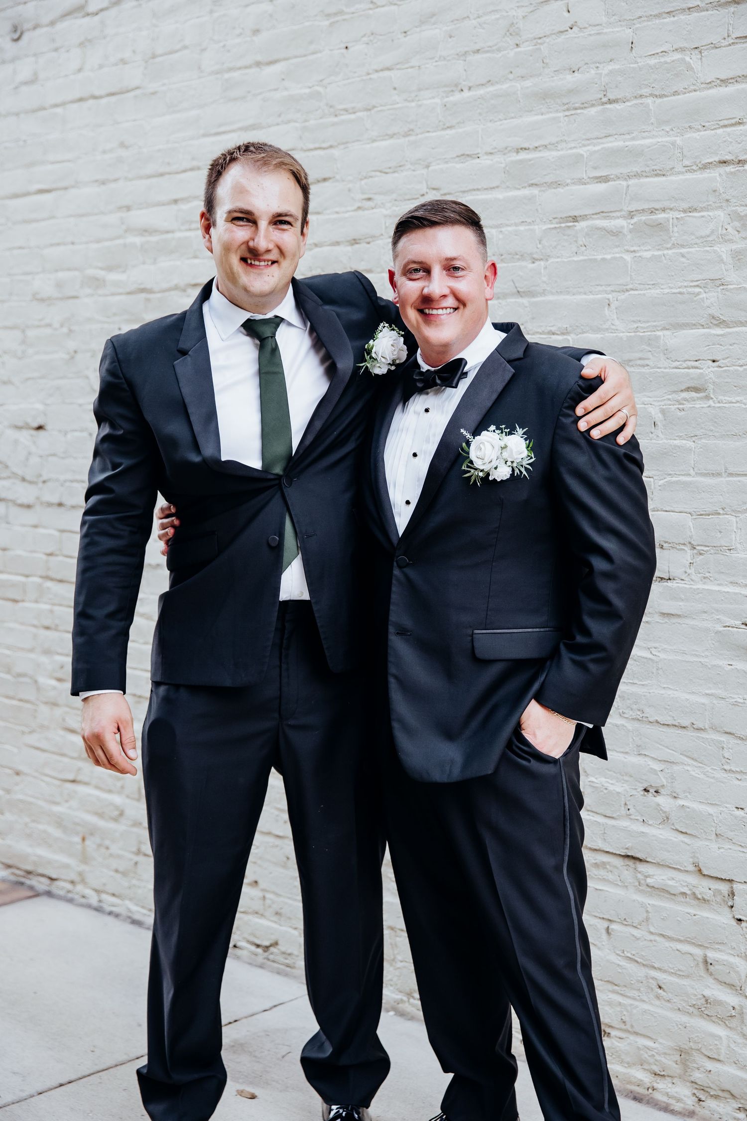 Groomsmen in black tuxedos pose together against white brick wall at wedding.