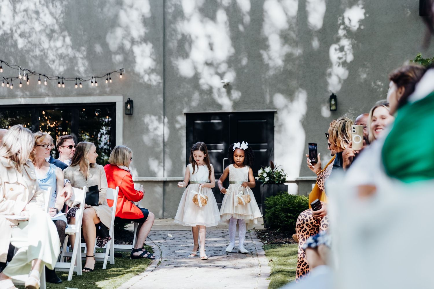 Wedding guests watch as flower girls walk down an outdoor ceremony aisle.