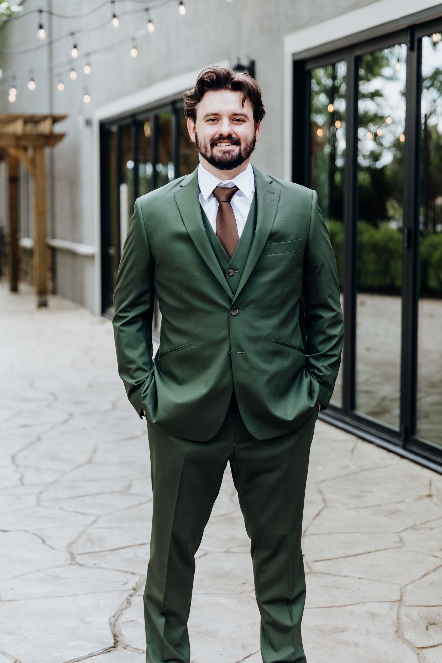 Groom wearing a dark green suit poses in an outdoor venue with string lights and stone patio.