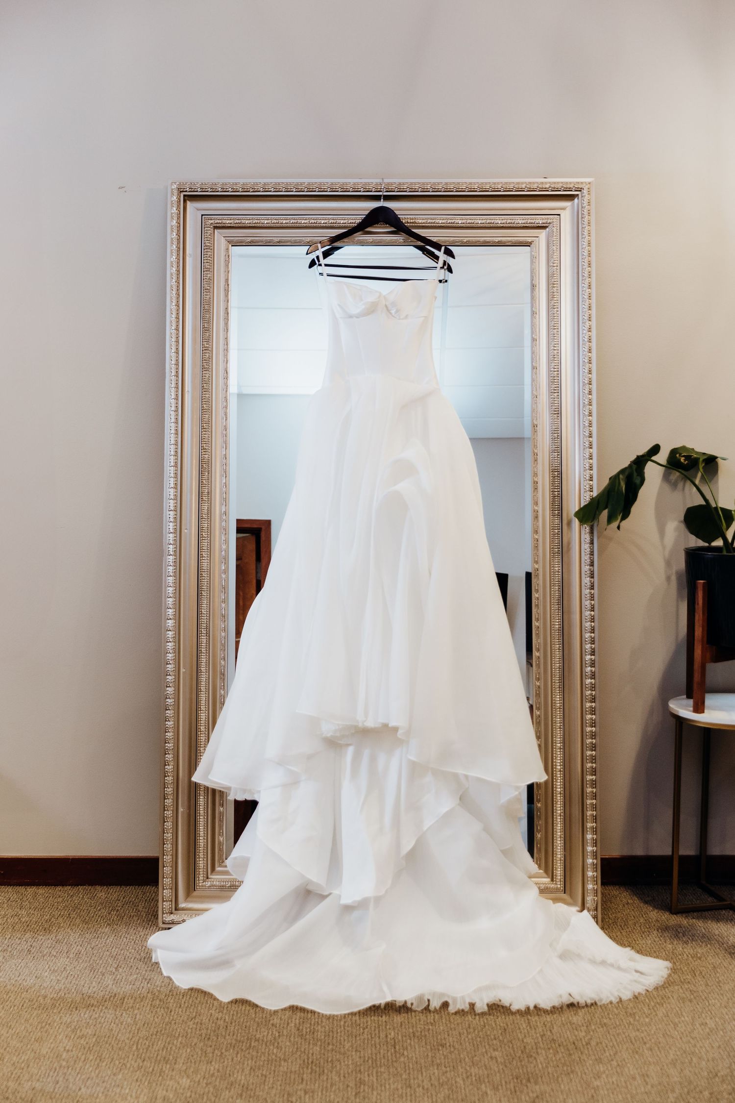 An elegant white wedding dress with a flowing train displayed on a hanger against an ornate framed mirror.