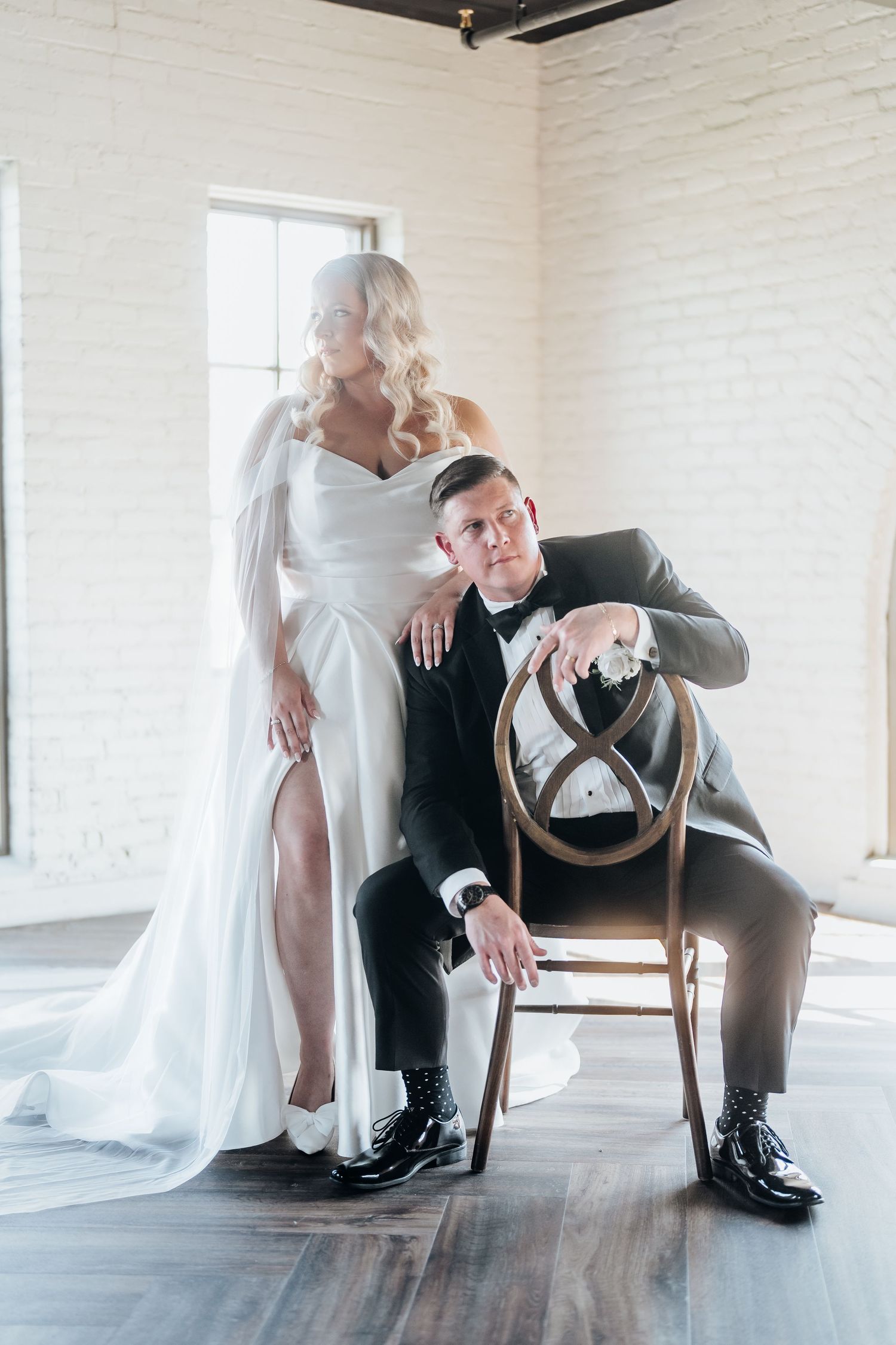 An artistic wedding portrait featuring a couple posing with a vintage chair in a minimalist white room.