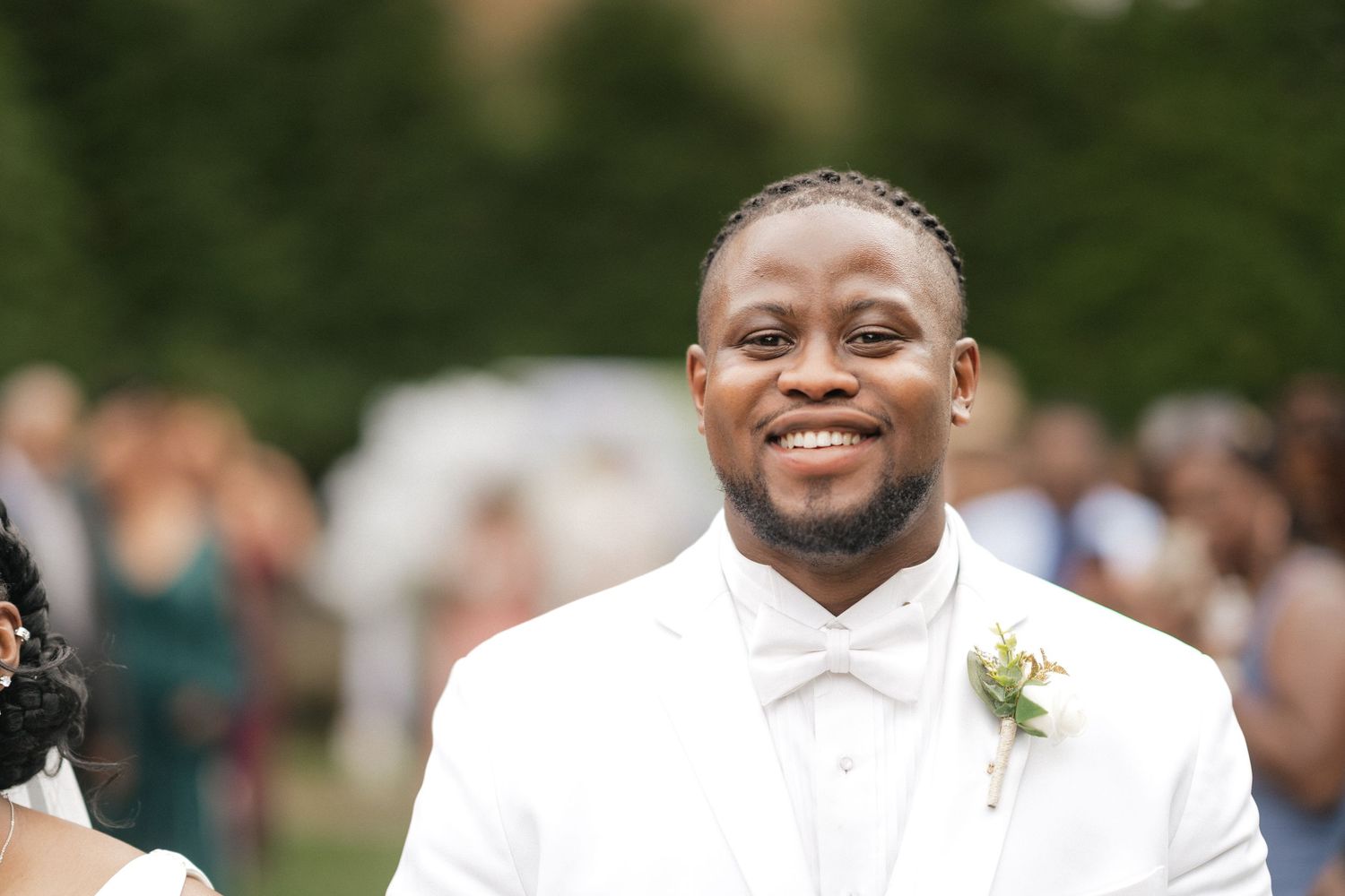 A joyful groom wearing a white tuxedo and bowtie smiles during an outdoor wedding ceremony.