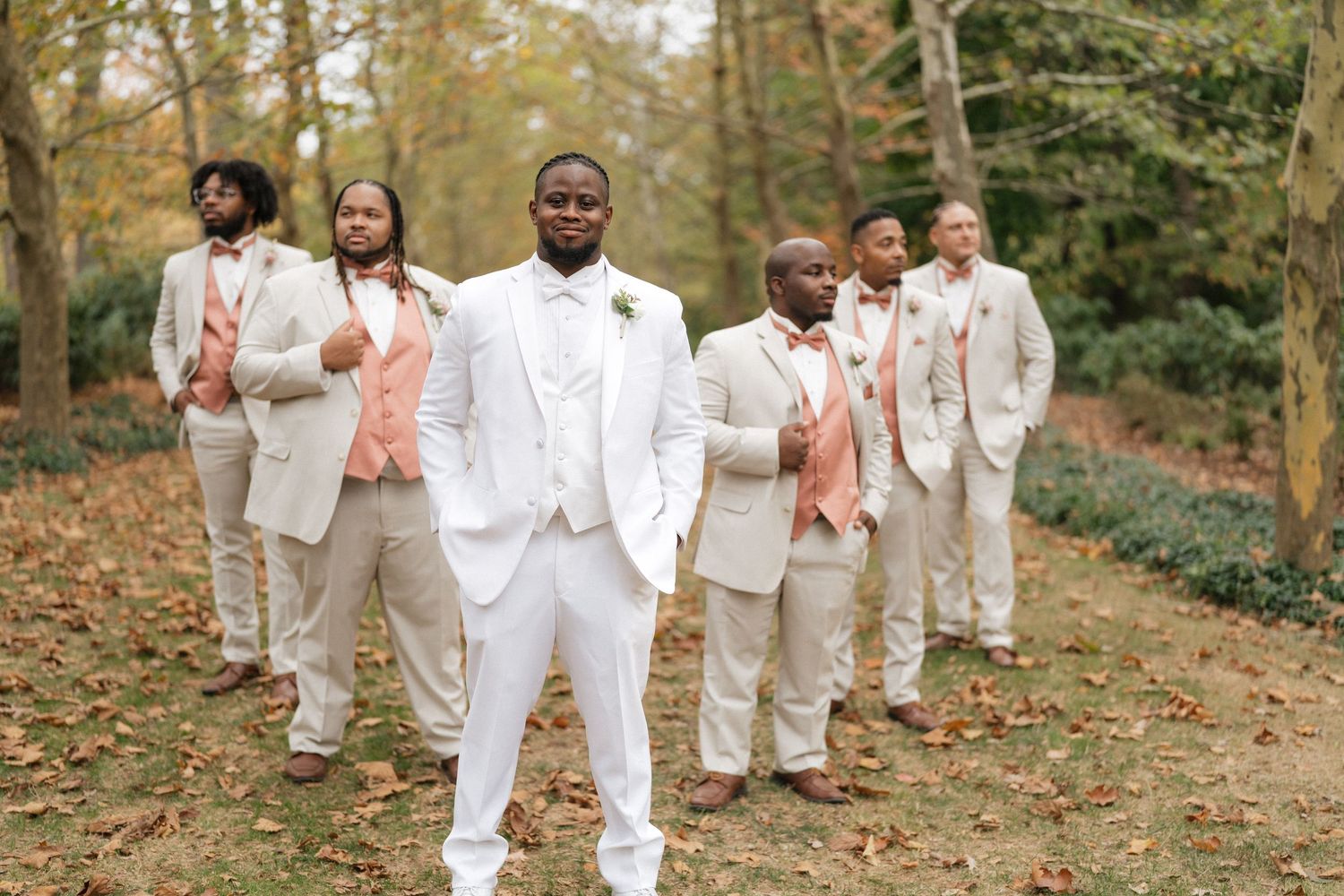 Wedding party members in cream-colored suits and dresses walk together on a leaf-covered autumn path.