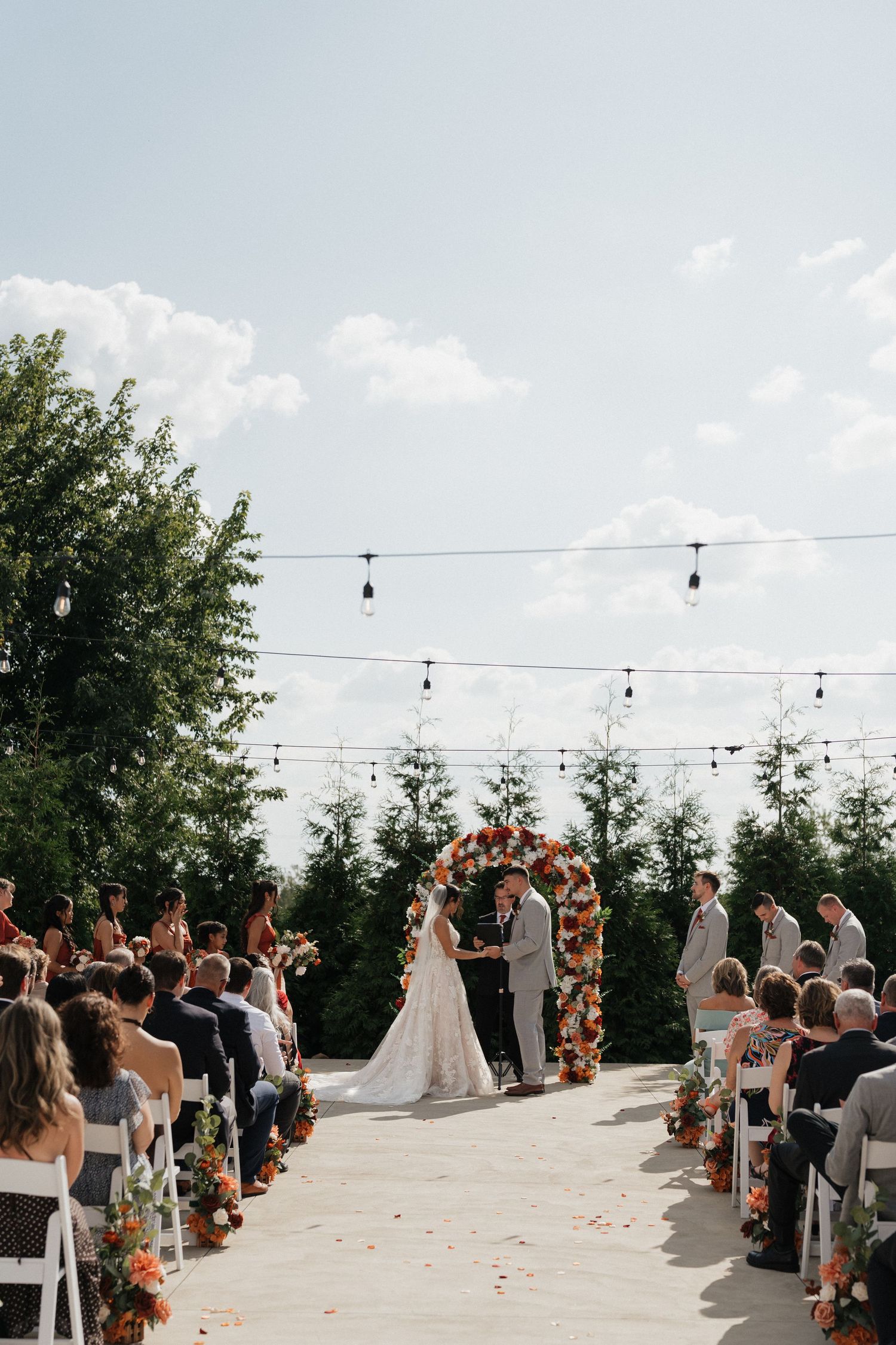 Outdoor wedding ceremony with string lights and floral arch overlooking a scenic view.