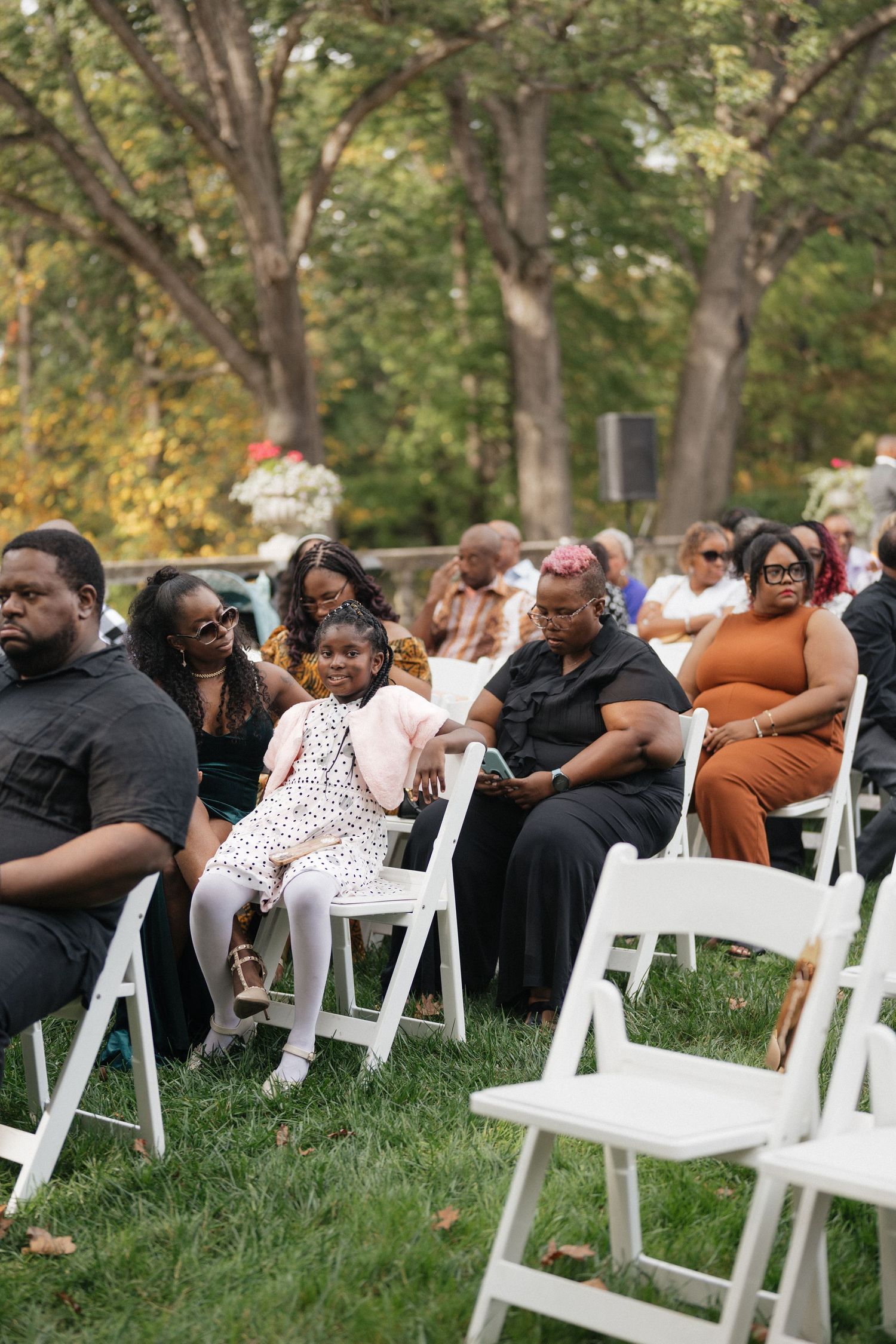 Wedding guests seated in white chairs on a grassy lawn during an outdoor ceremony.