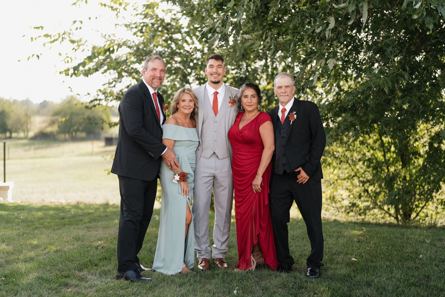 Wedding party in gray suits and red dresses pose together under trees.