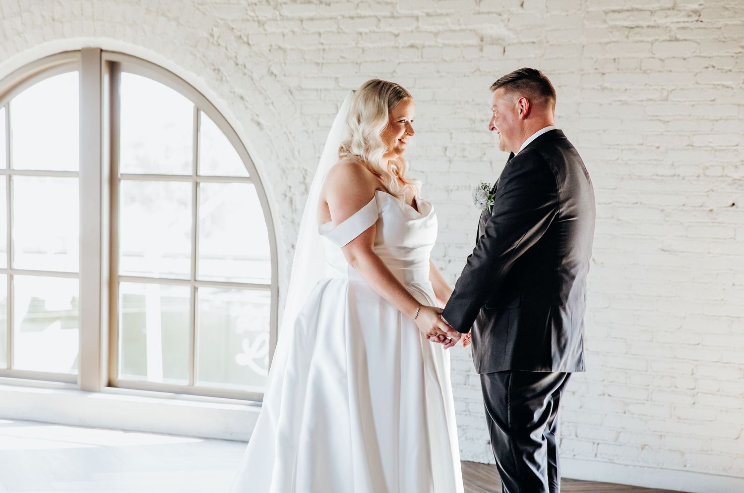 Elegant first look wedding moment in a bright white brick room with arched windows.