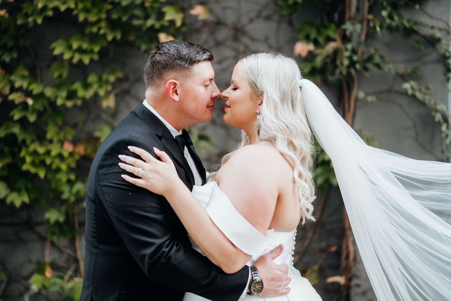 Romantic wedding portrait of couple embracing against ivy-covered wall.