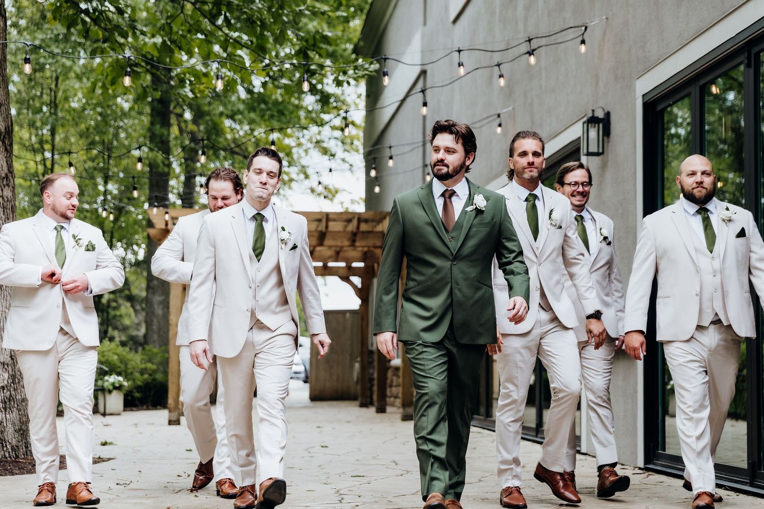 Groomsmen in light gray suits and one in dark green walk together outdoors.