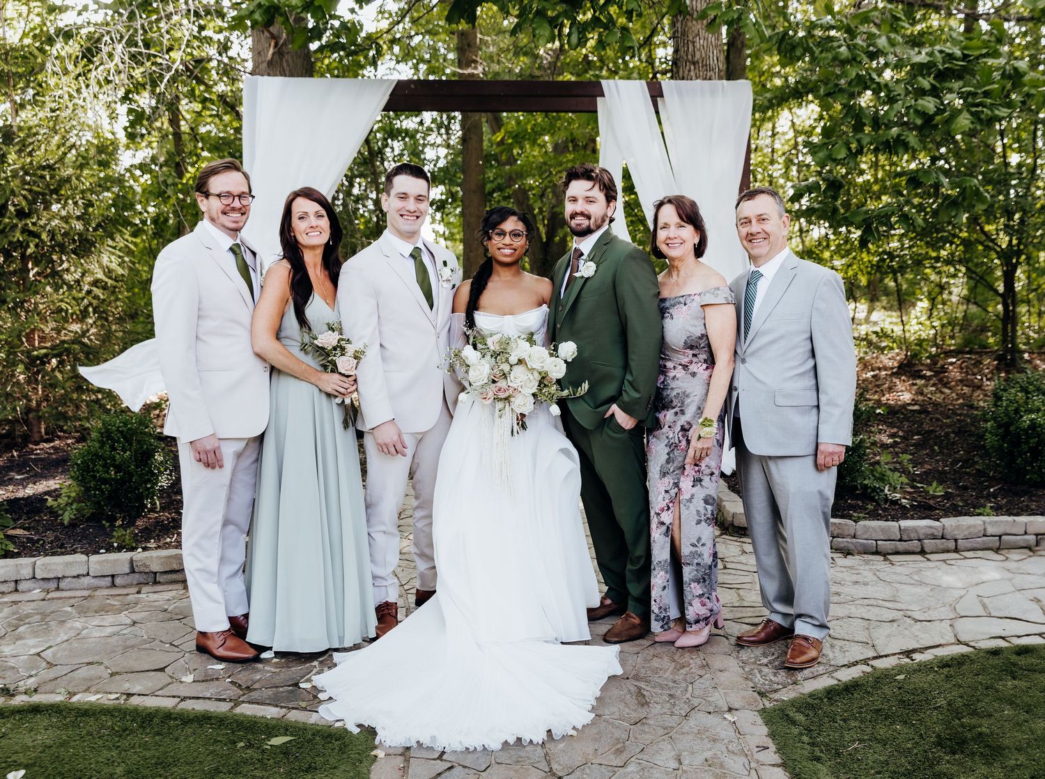 Wedding party poses together under wooden arch with mix of white suits and formal attire.