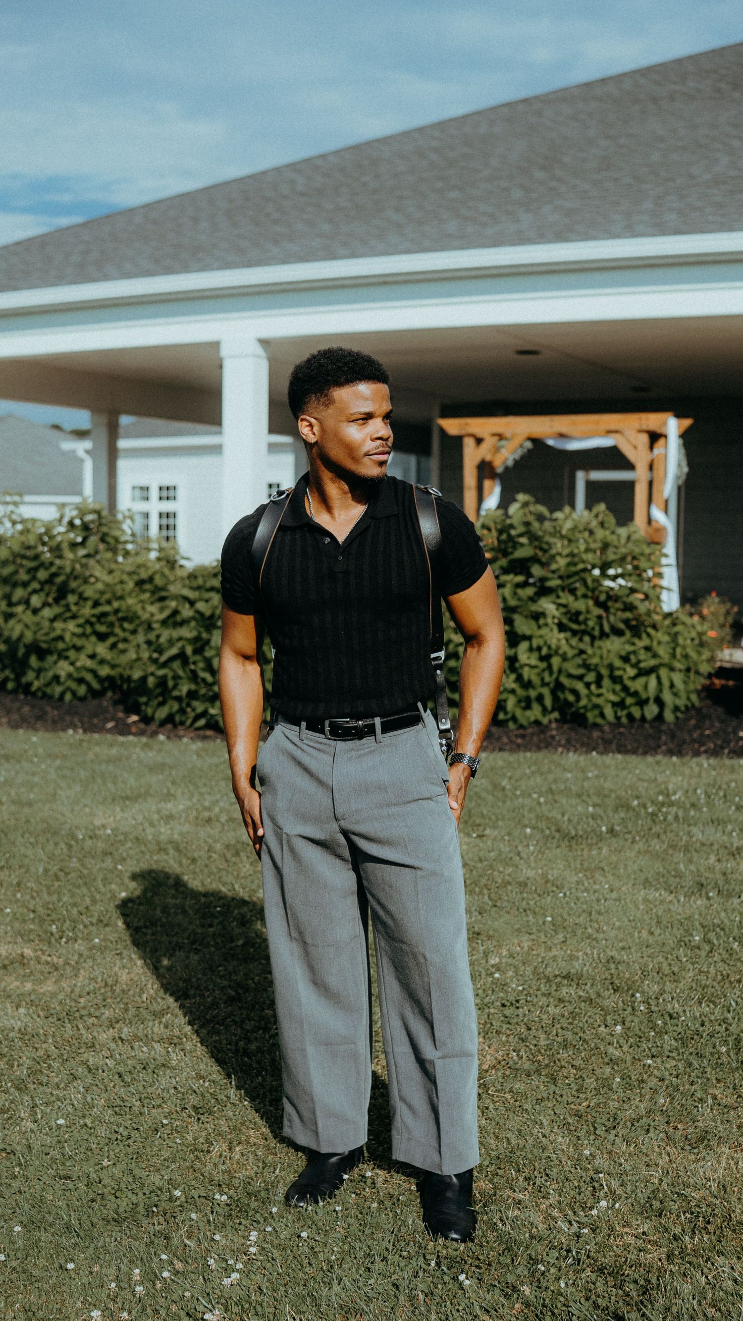 Person in a black shirt and gray pants stands on grass in front of a house with white trim.