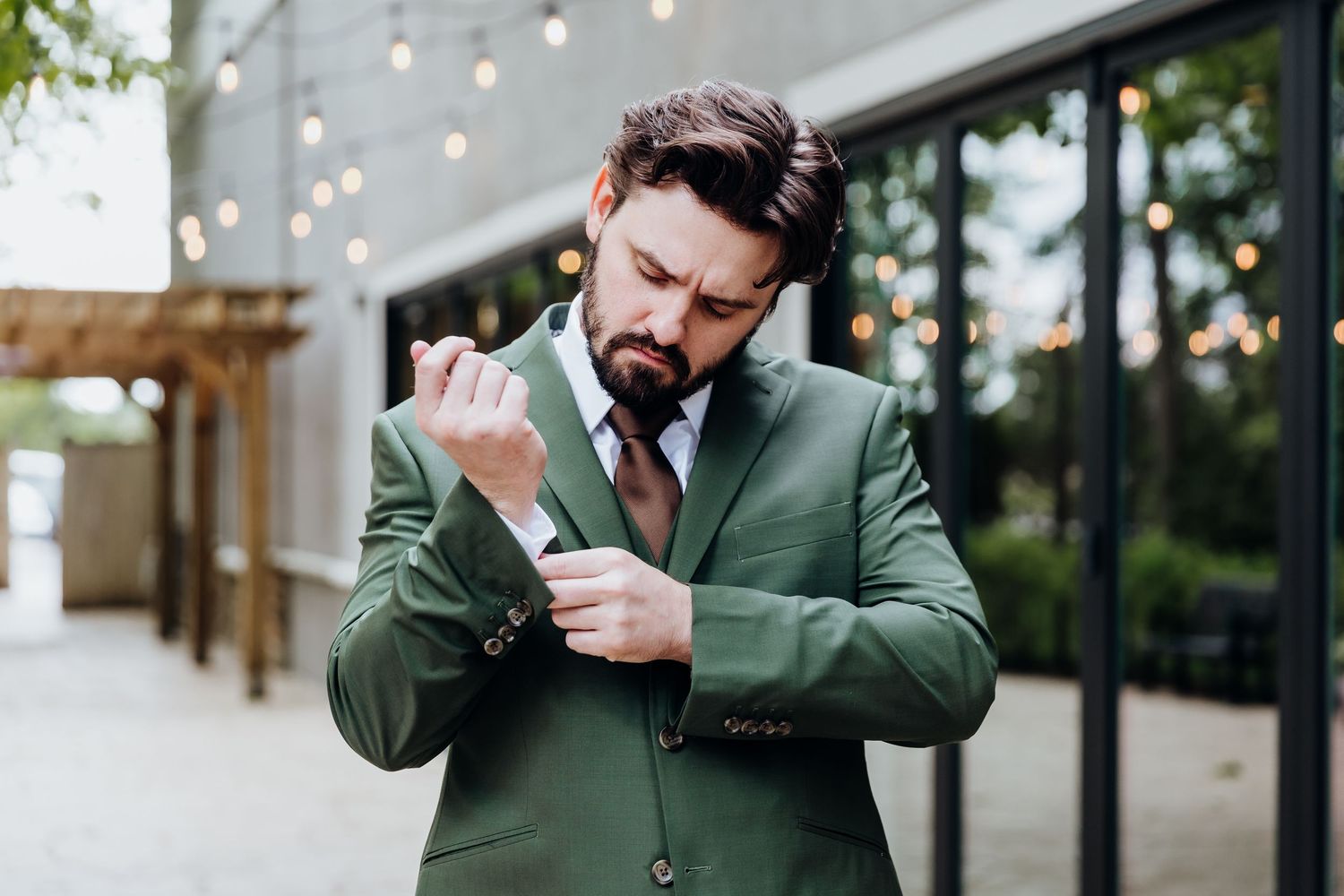 A person in a green suit adjusts their sleeve cuff while standing outside a building with string lights.