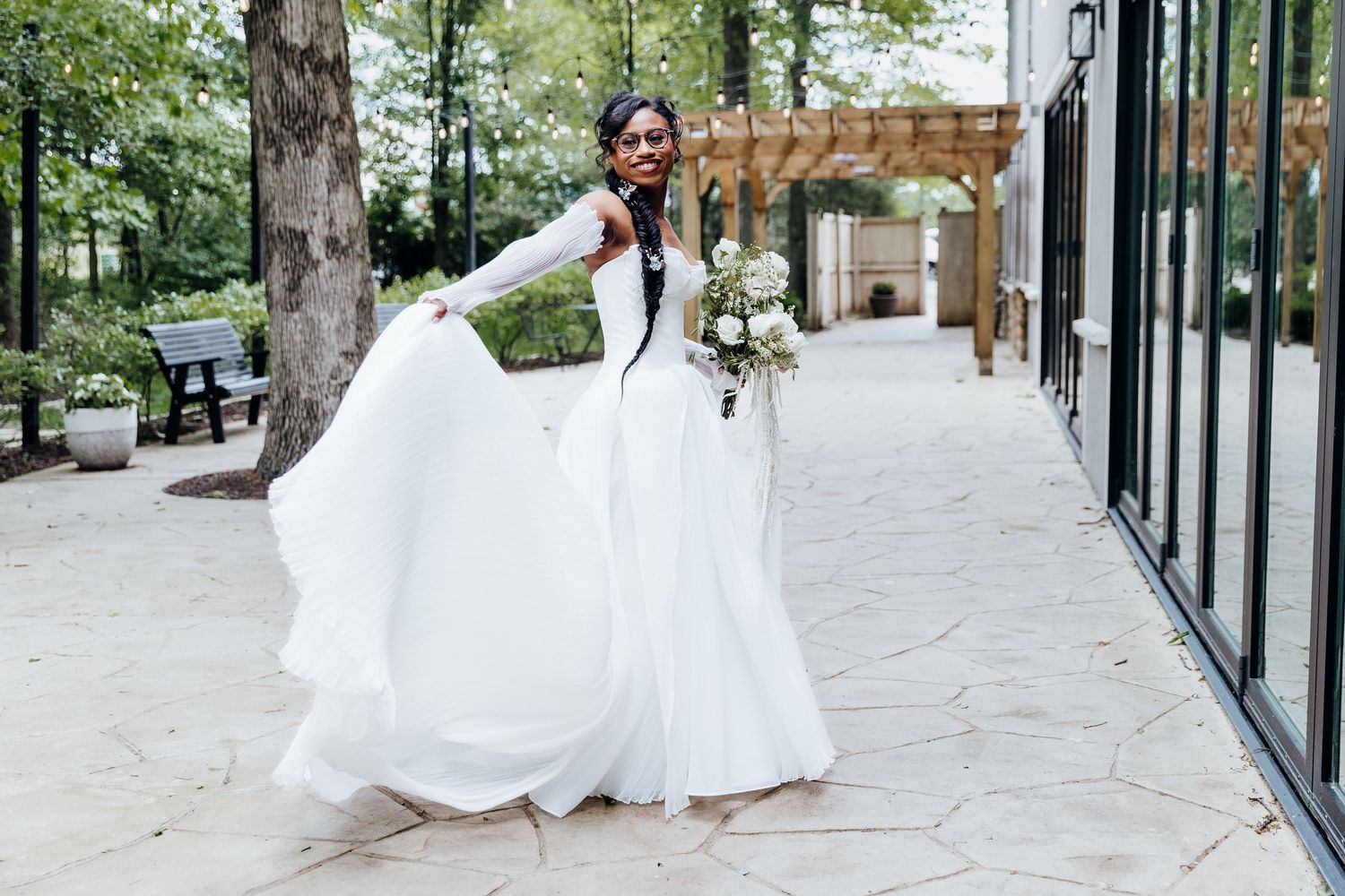 A bride in a flowing white gown twirls on a stone patio with a garden pergola in the background.