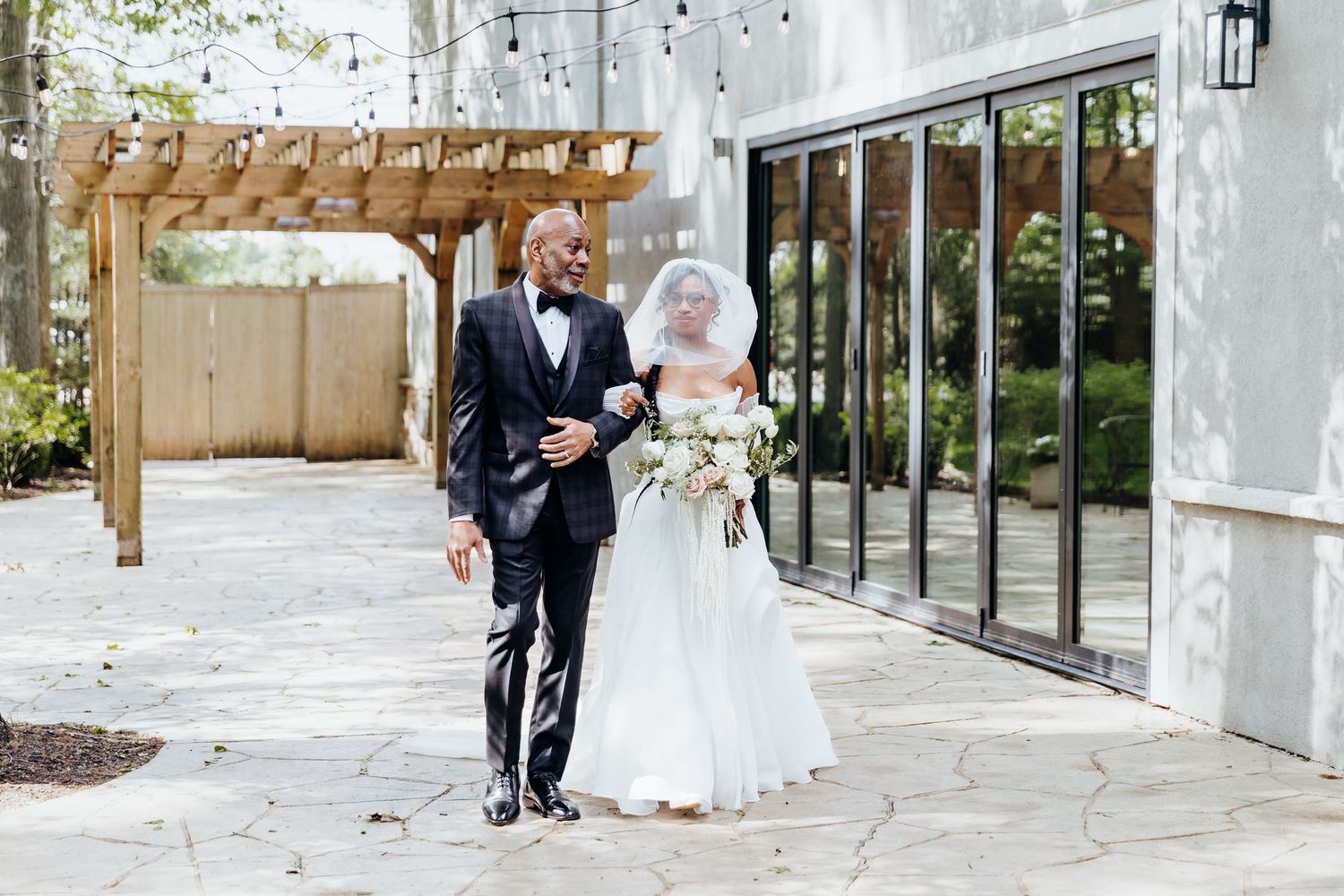A couple exits their wedding ceremony under a wooden pergola near modern glass doors.