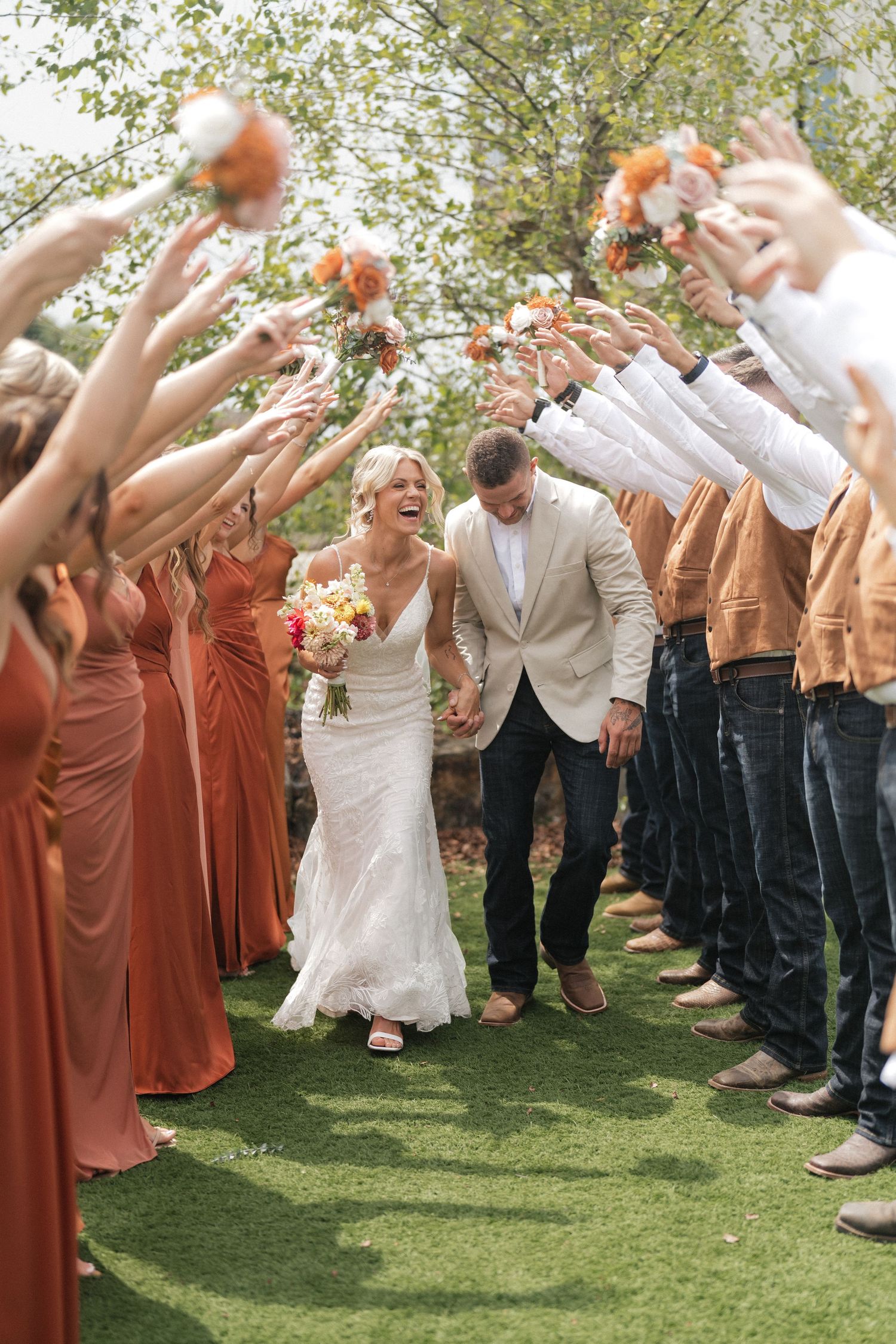 Wedding party celebrates with guests in rust-colored attire throwing confetti on a sunny lawn during outdoor reception.