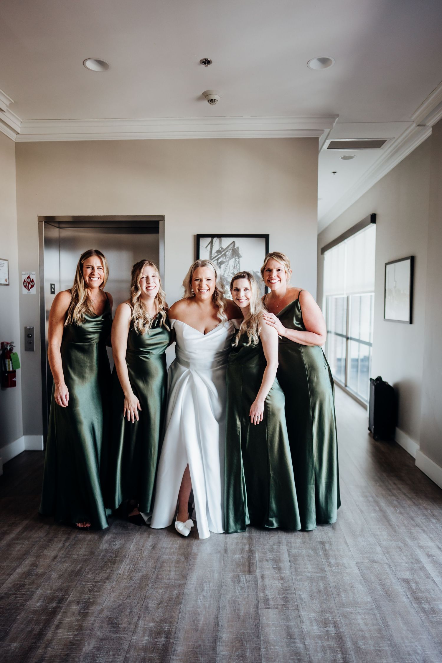 Wedding party in sage green bridesmaid dresses and white bridal gown posing playfully in a hallway.