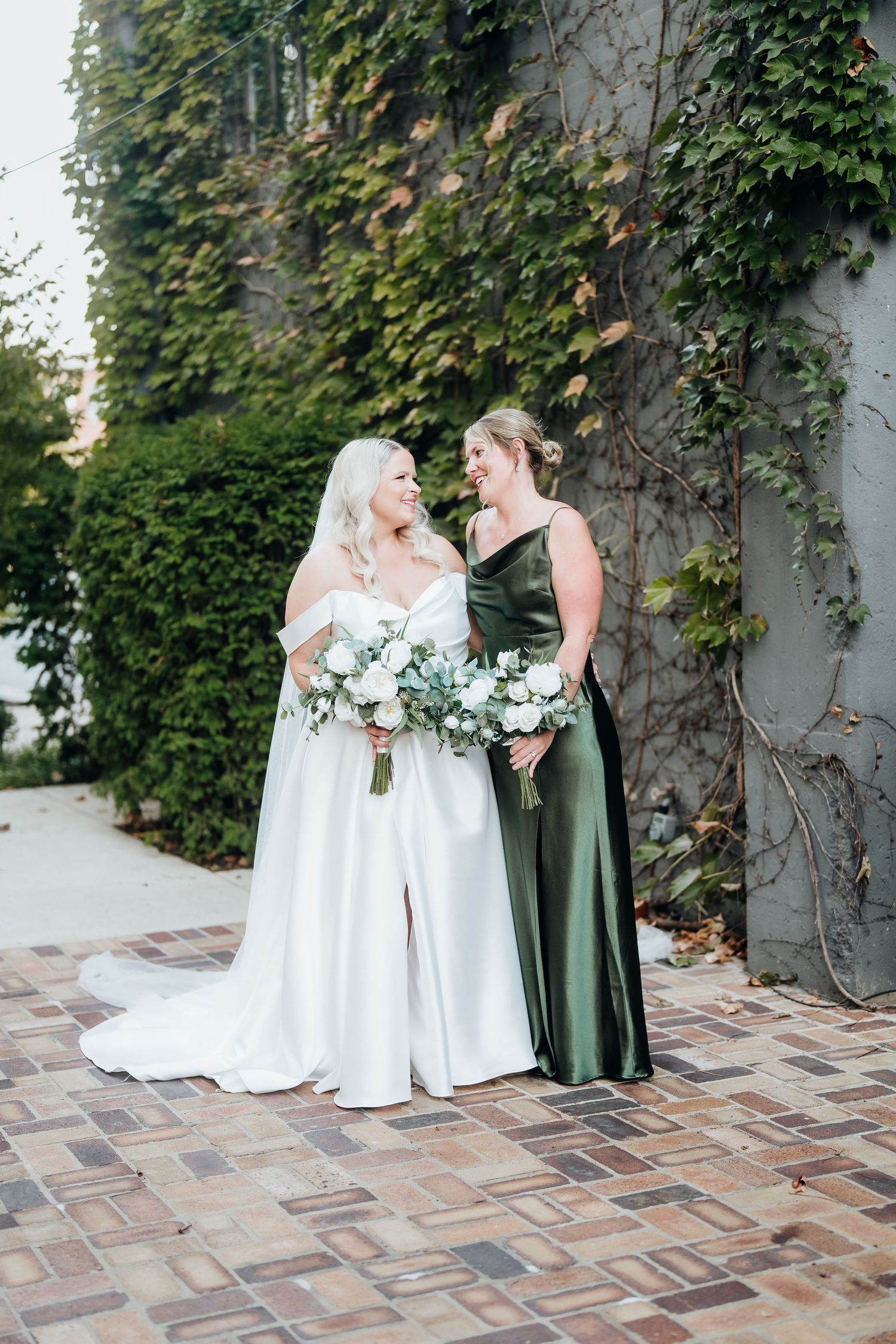 Two people in wedding attire share a moment on a stone patio with ivy-covered walls.