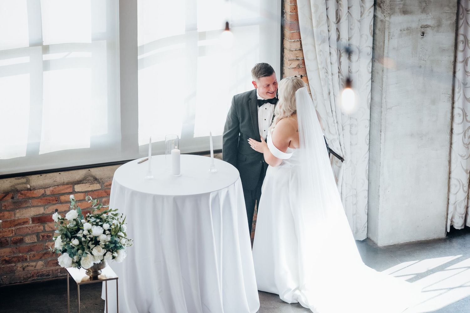 Intimate wedding moment captured in a bright sunlit room with white decor and floral arrangements.