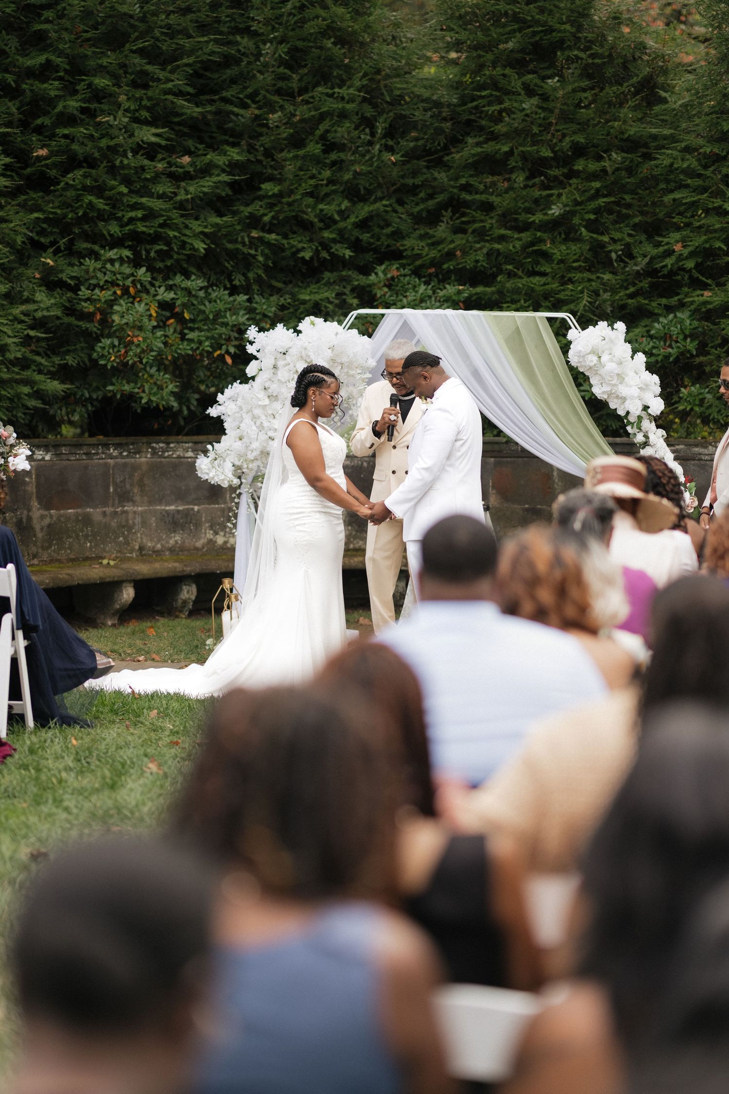 Outdoor wedding ceremony with white draped arch and guests seated on lawn.