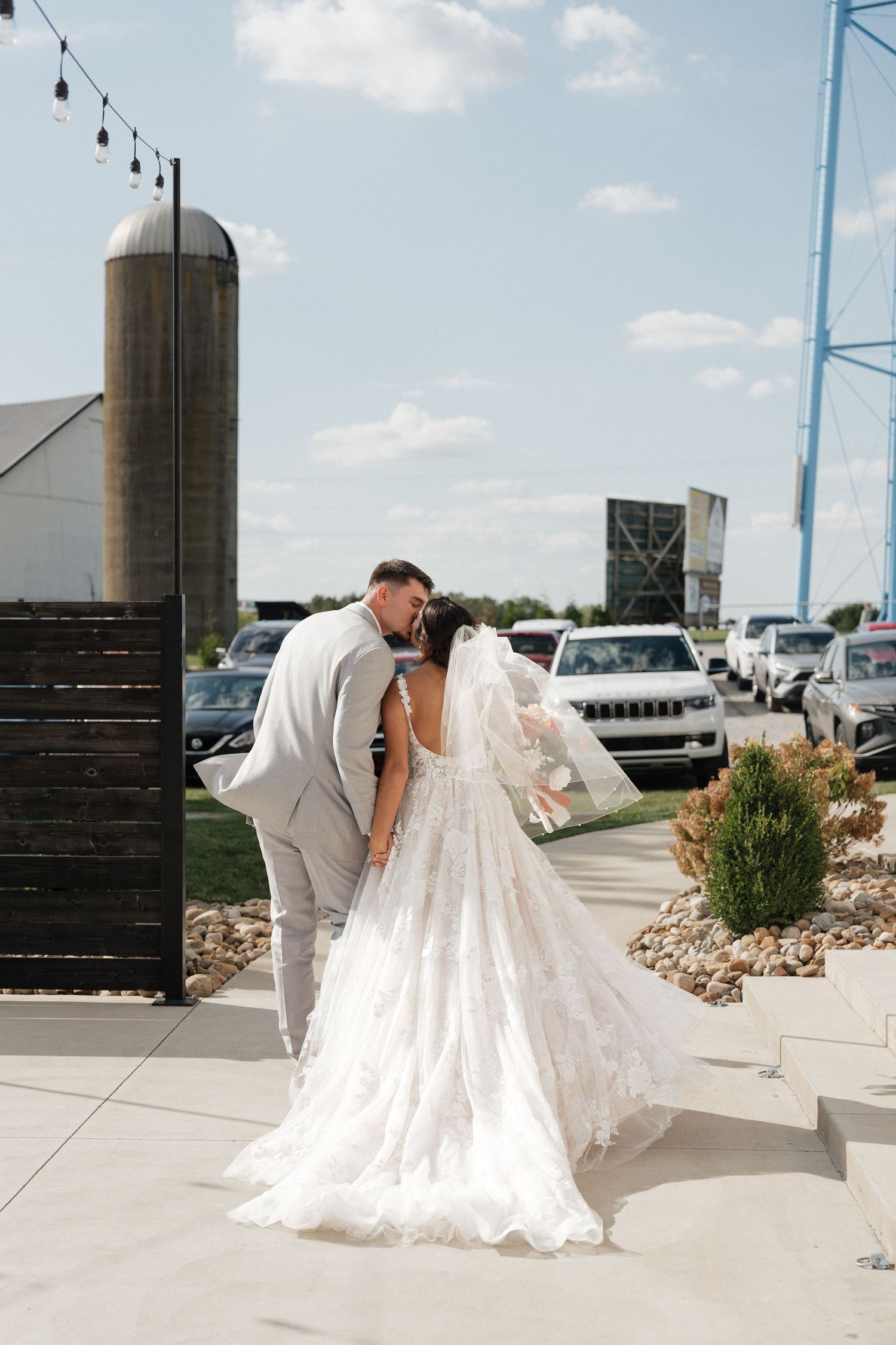 Wedding couple walking together near a rustic silo and parked cars.