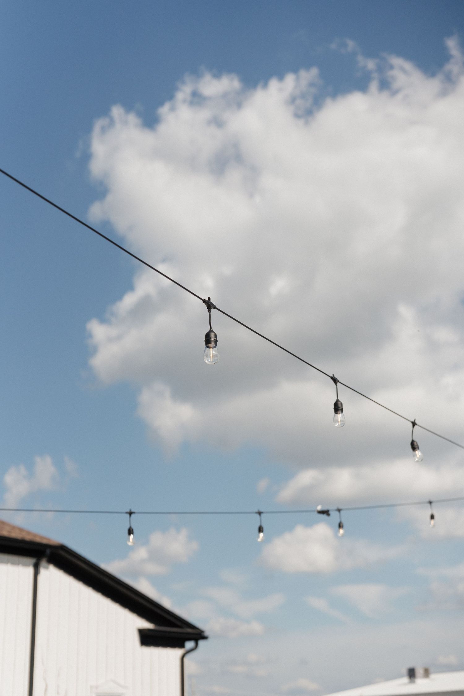 String lights hang against a bright blue sky with puffy white clouds and a white barn in background.