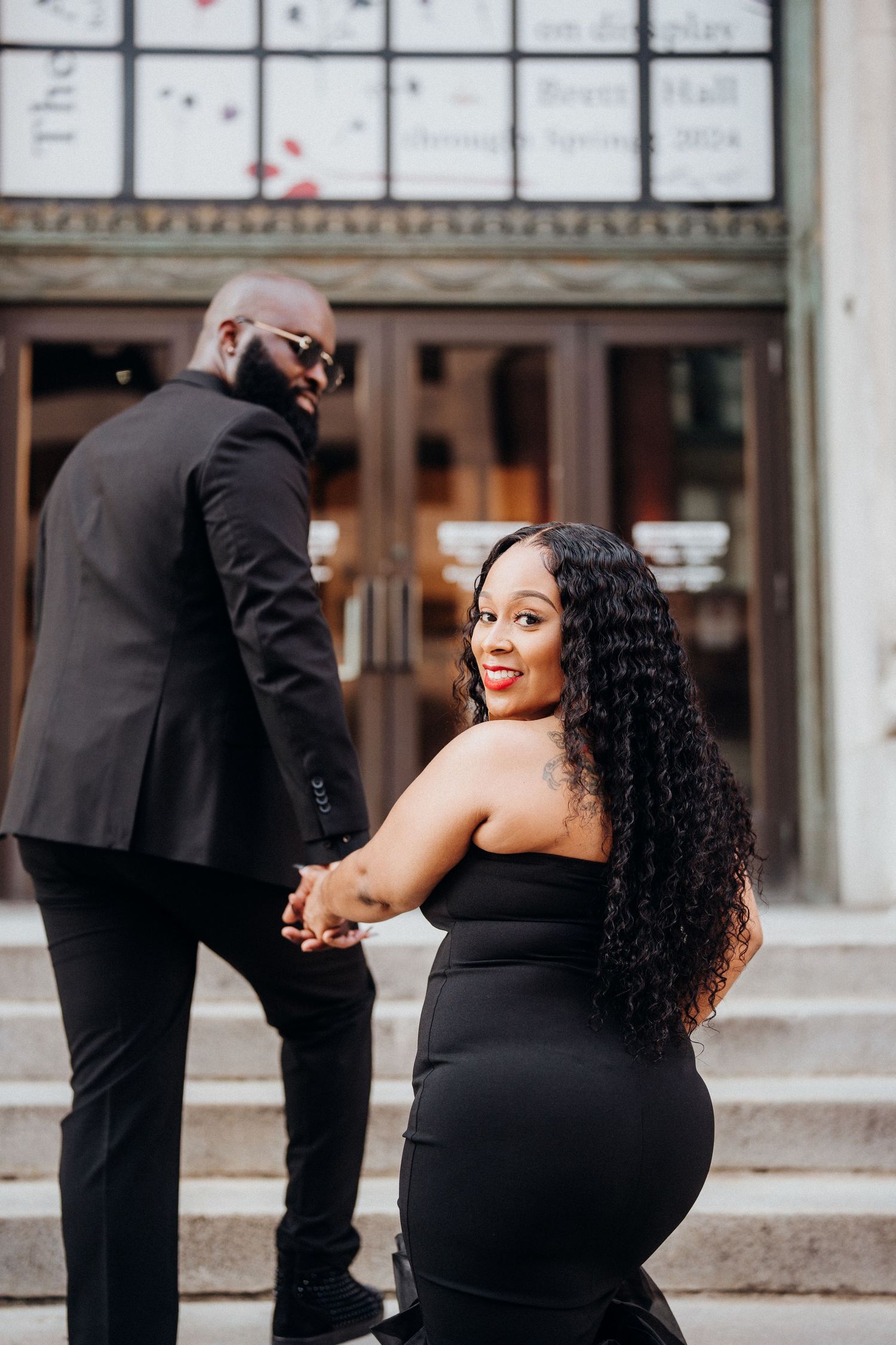A couple in matching black outfits poses on stone steps in front of a building.