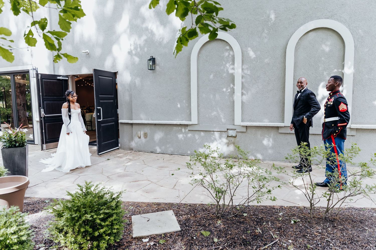 Wedding party poses in front of a modern building with white walls and green foliage.