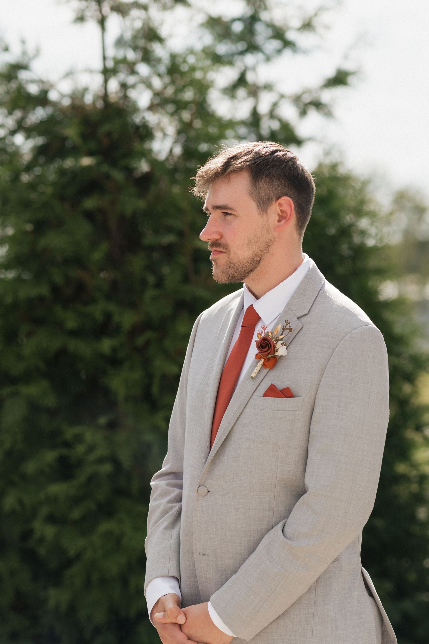 Groom in light grey suit with rust-colored tie standing during outdoor wedding ceremony.