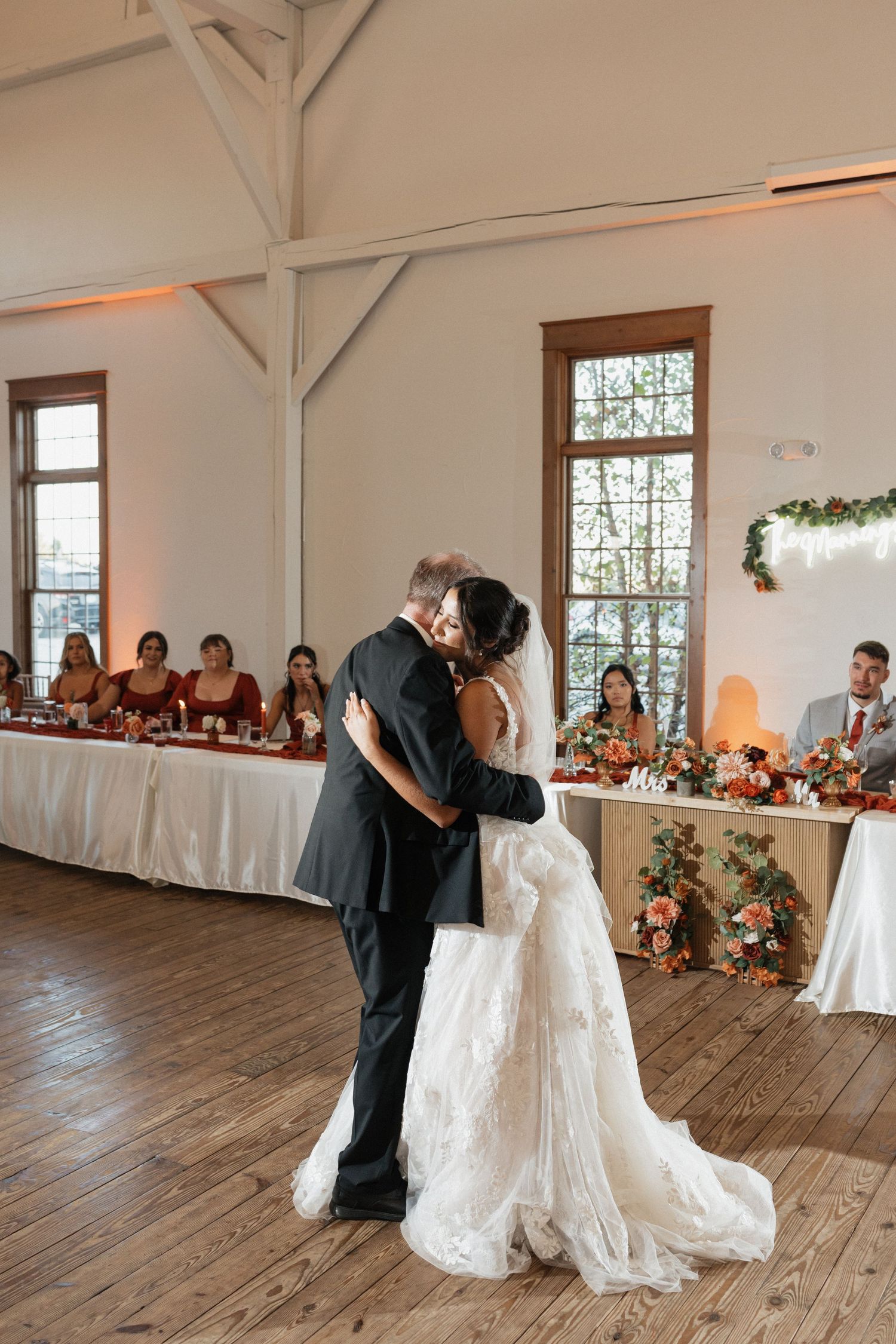 Newlyweds share their first dance on a wooden floor in a bright reception venue with floral decorations.
