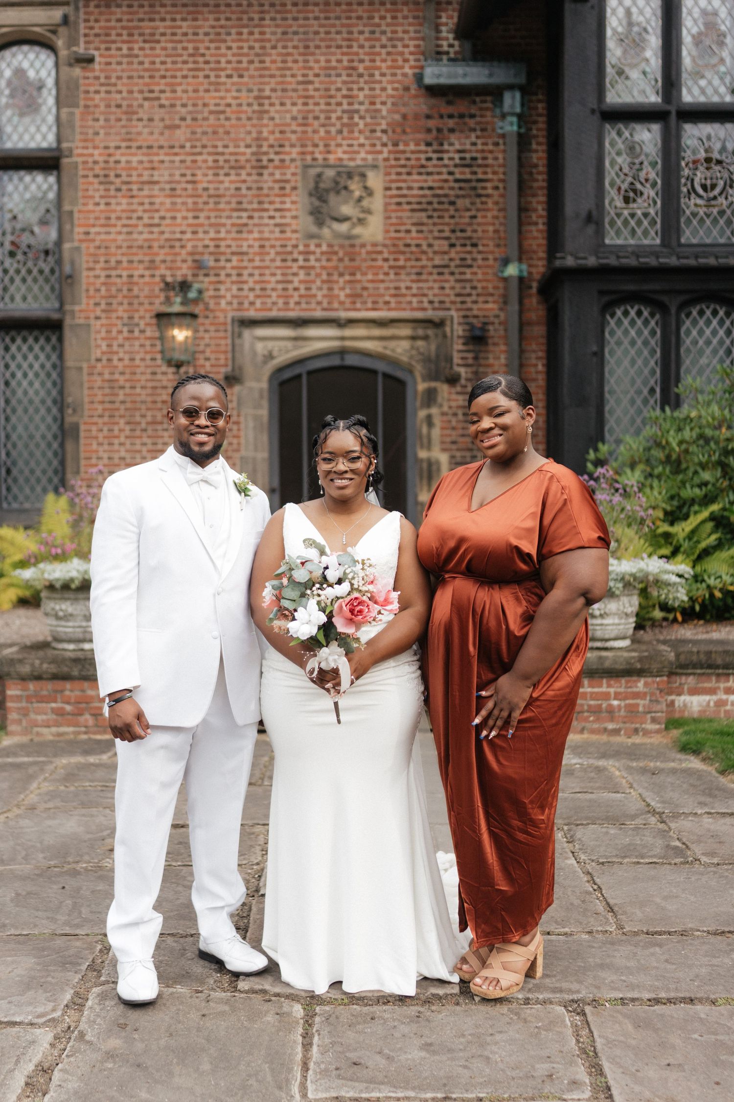 A wedding party poses in front of a historic brick building with the couple in white and guest in rust-colored dress.