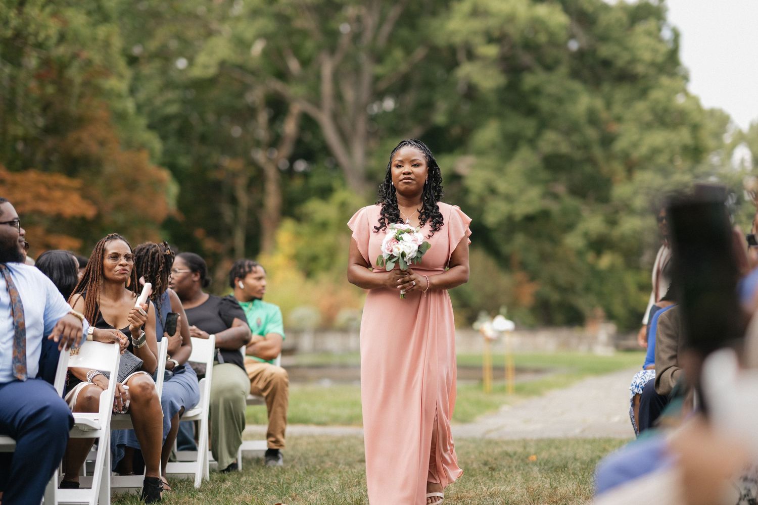Wedding ceremony guests seated in rows outdoors with bridesmaids in coral dresses.