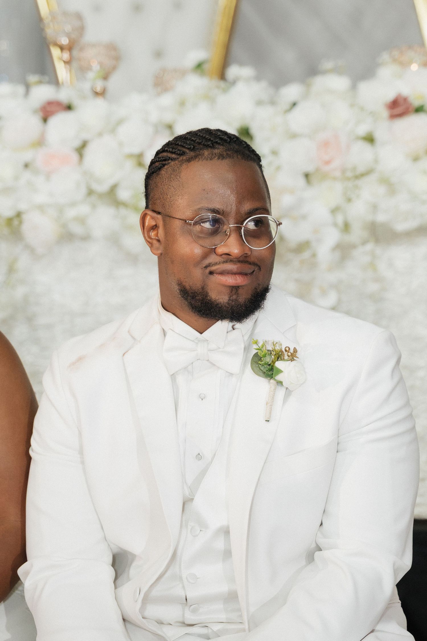 Wedding party member in crisp white suit and glasses poses against floral backdrop.