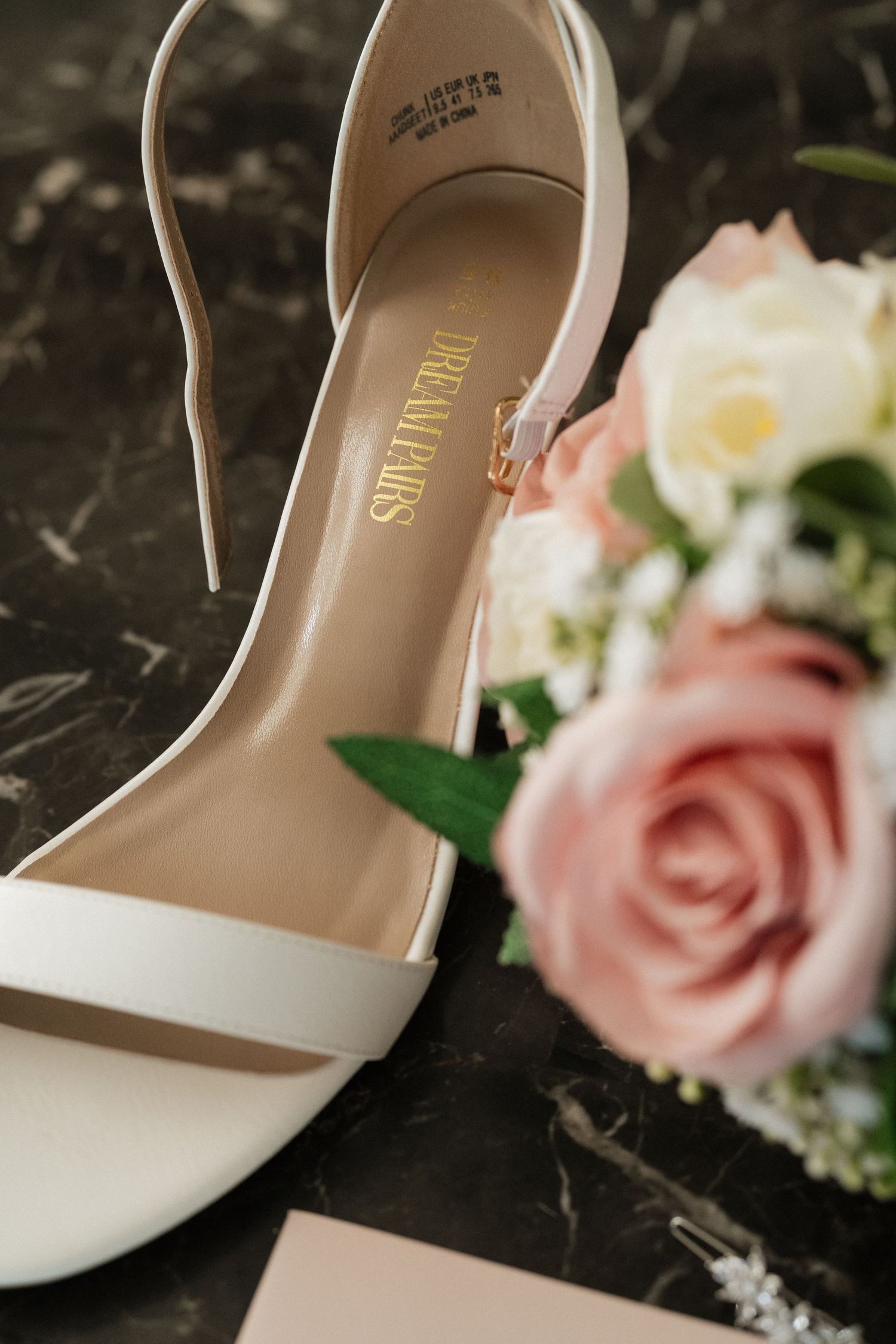 White bridal shoe next to pink roses on dark marble surface.