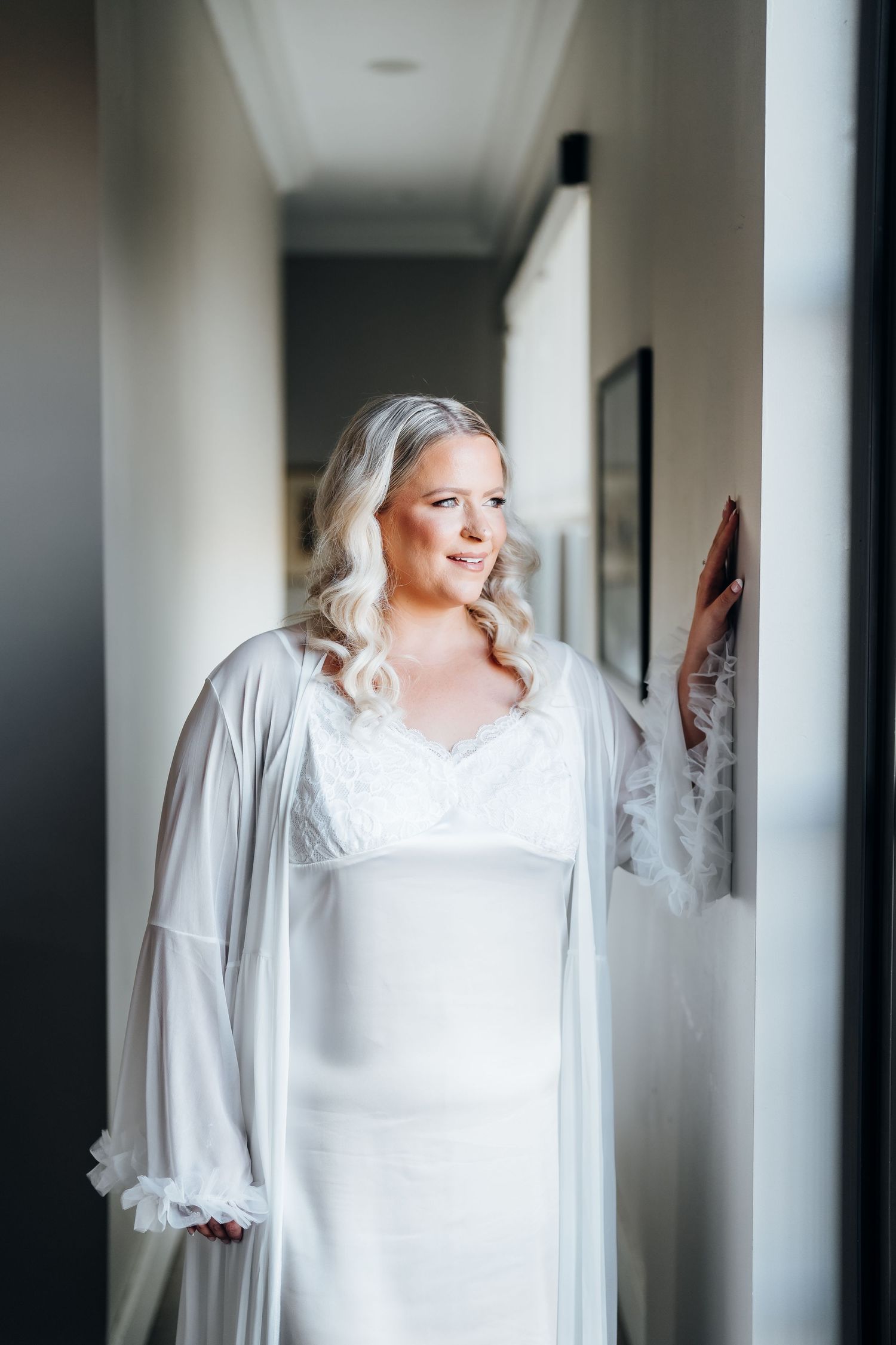 Person wearing an elegant white dress poses in a sunlit corridor.