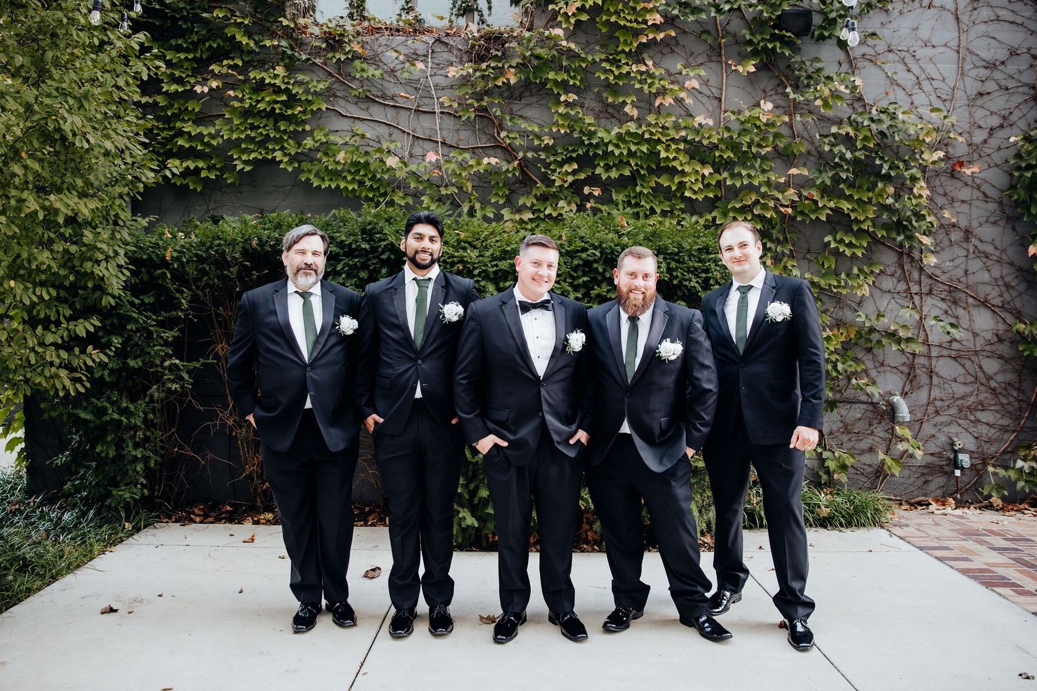 Groomsmen in black tuxedos pose together against a brick wall with greenery.