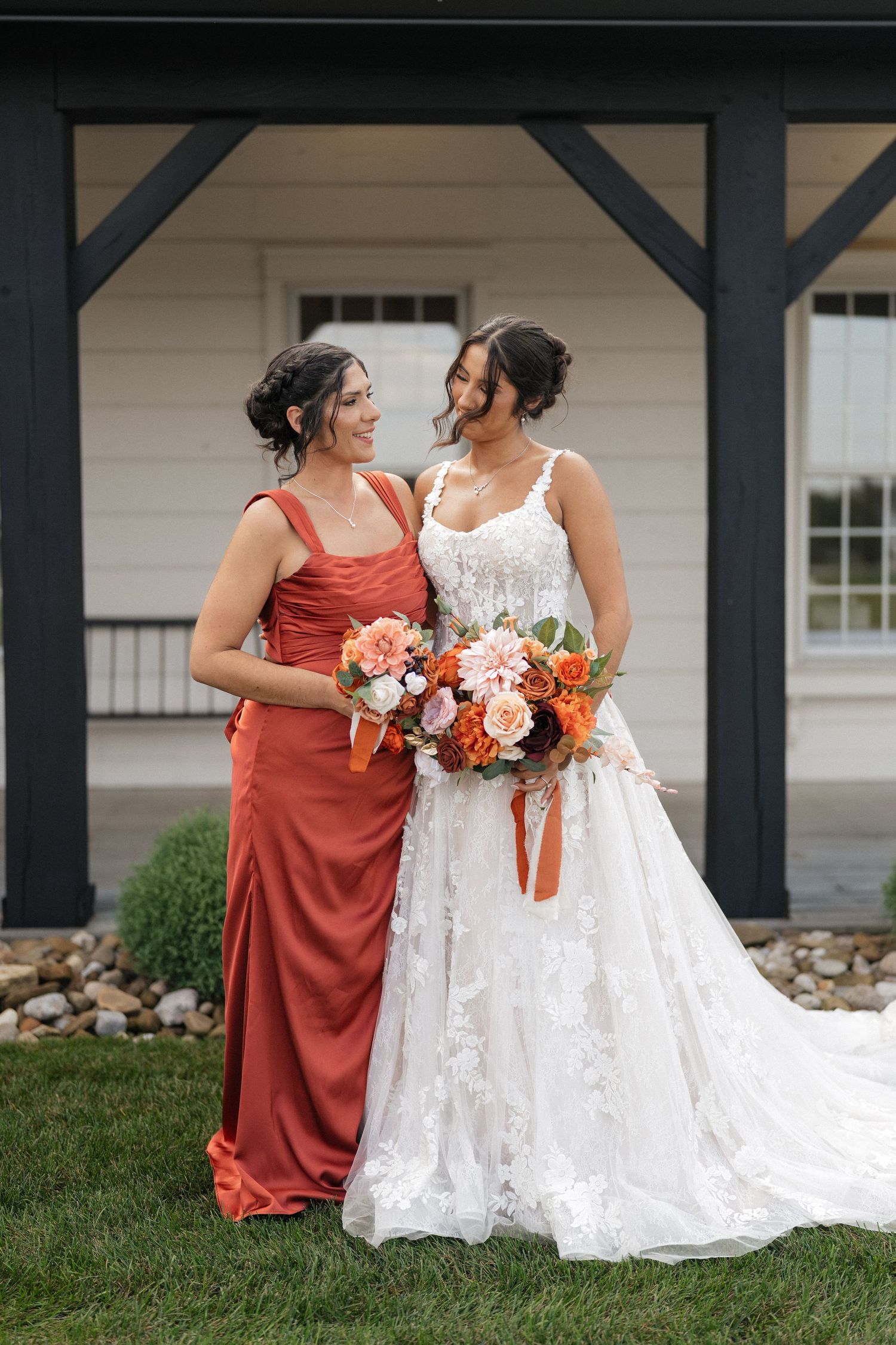 Wedding party in rust-colored dresses and white lace gown posed outdoors by rustic building.